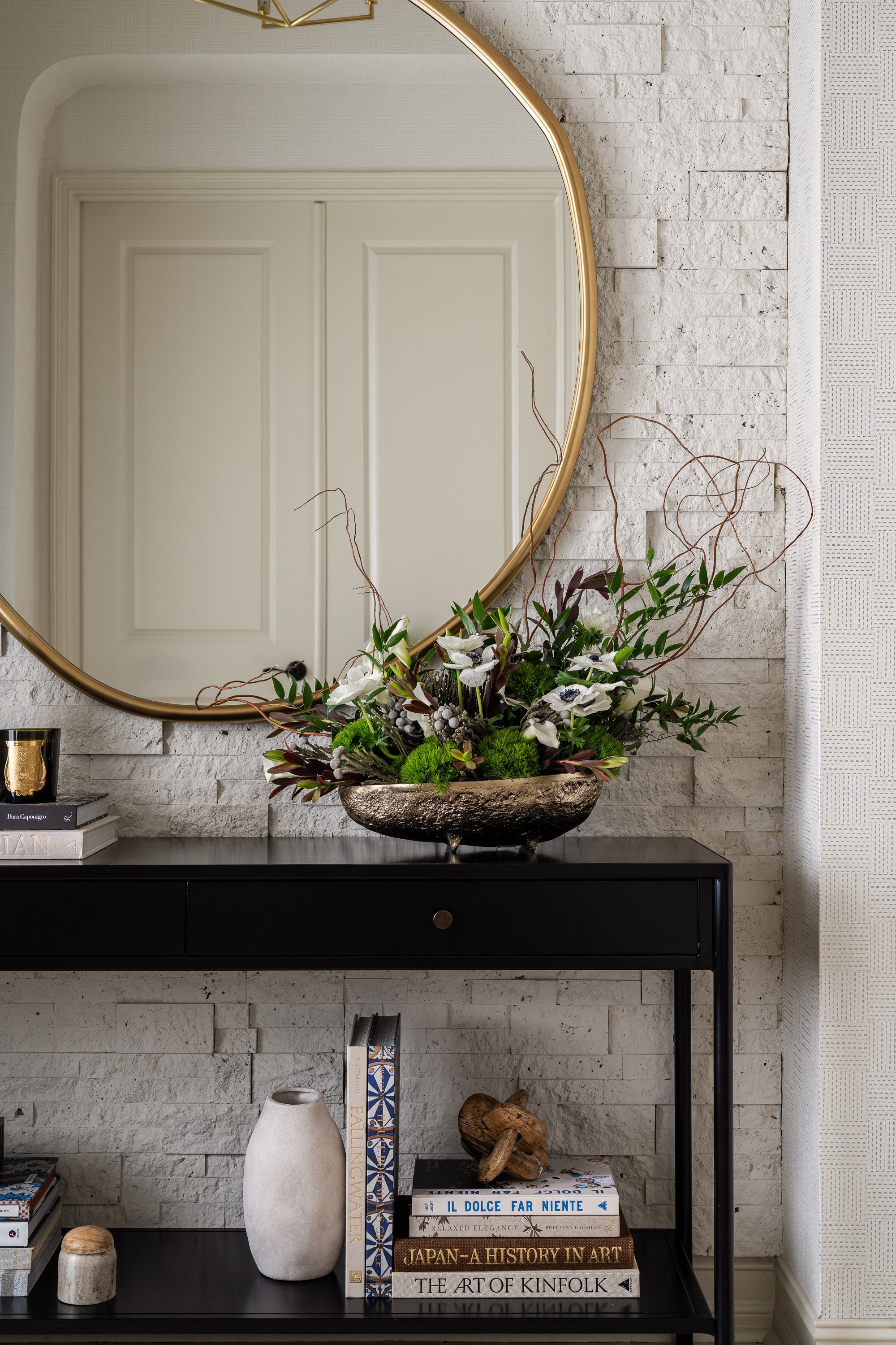 A decorative arrangement of greenery and white flowers in a metallic bowl sits on a black console table below a gold-rimmed mirror, with books and a vase on the shelf below.