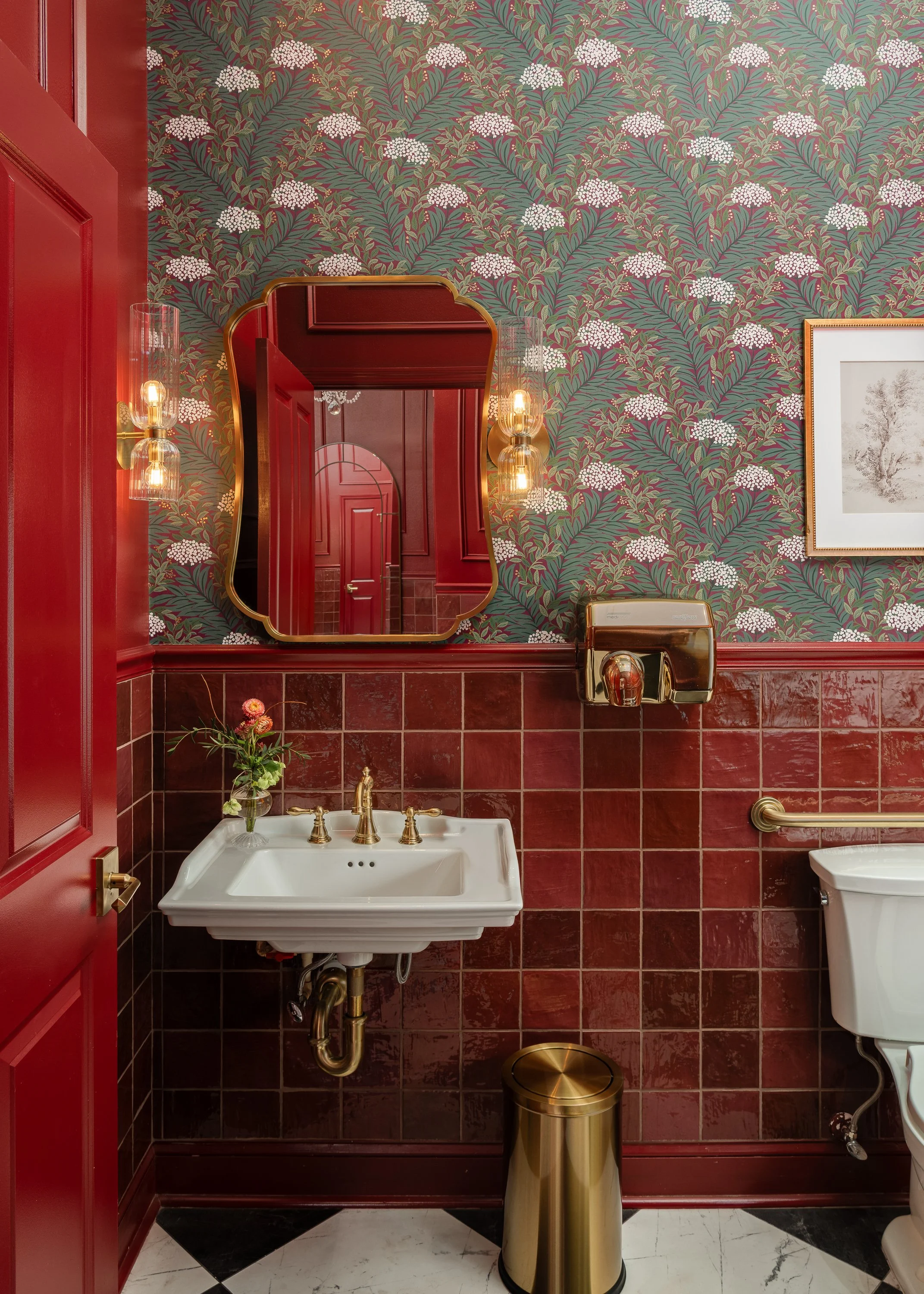 A commercial restroom design with red tiled walls, floral wallpaper, a white sink with gold fixtures, a gold-framed mirror, and a toilet.