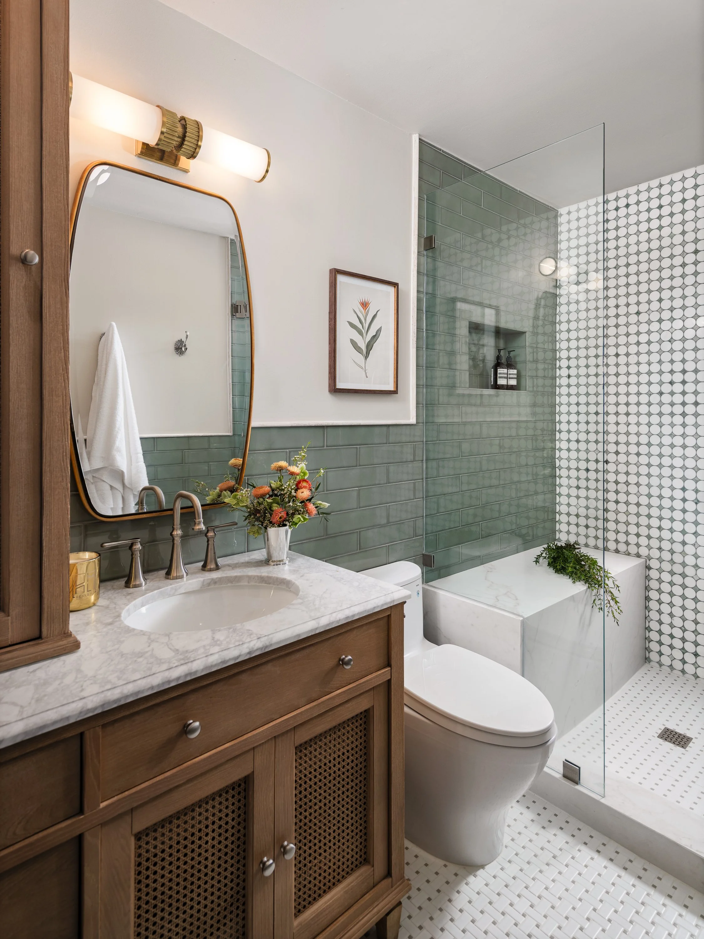 A modern bathroom features a wooden vanity with a marble countertop, a white sink, and a gold-framed mirror. A green tiled shower with a glass enclosure and a built-in bench is visible, along with a white toilet and a small framed botanical print.