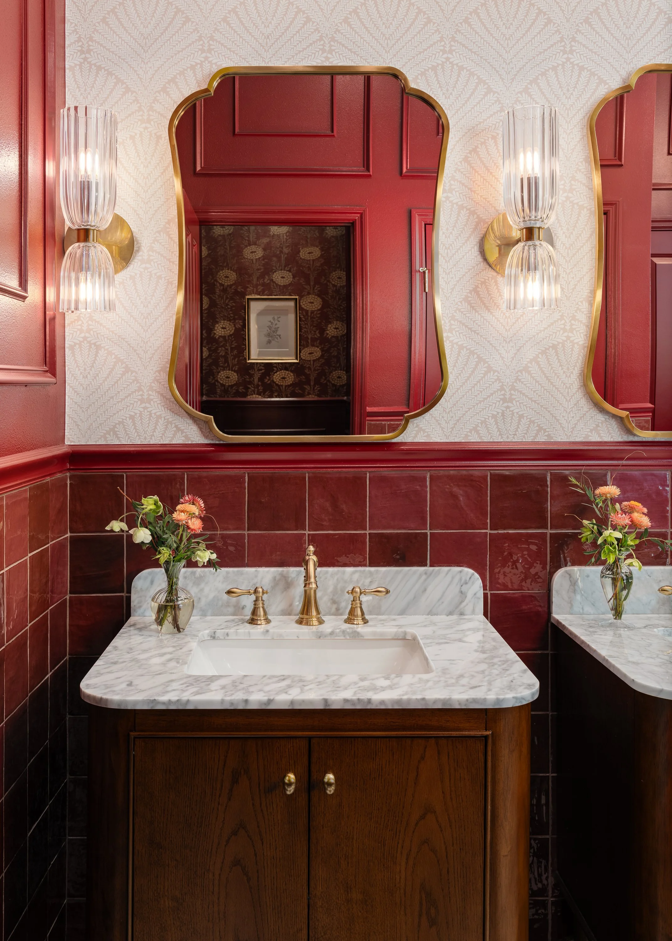 A commercial restroom design vanity with a marble countertop, brass fixtures, and a bouquet of flowers in a vase. The walls are tiled in deep red and feature a decorative wallpaper and a large ornate mirror.