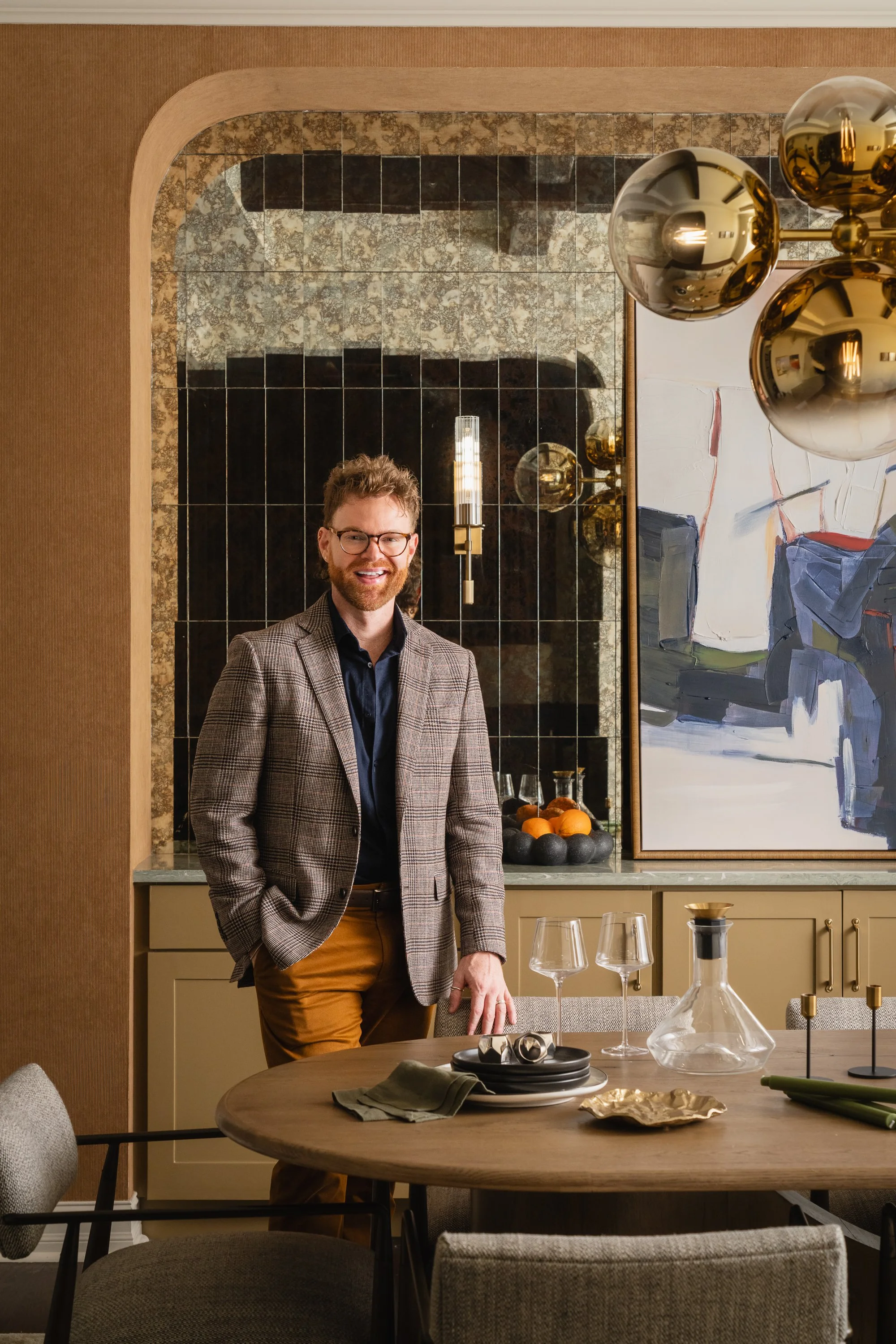 A man with glasses and a beard wearing a checked blazer, black shirt, and brown pants standing in a modern dining room. The room features a round dining table set with glasses, plates, and decorative items. Behind him, there is a kitchen area with a black and gold tiled backsplash, an abstract painting, and gold-colored spherical pendant lights hanging from the ceiling.