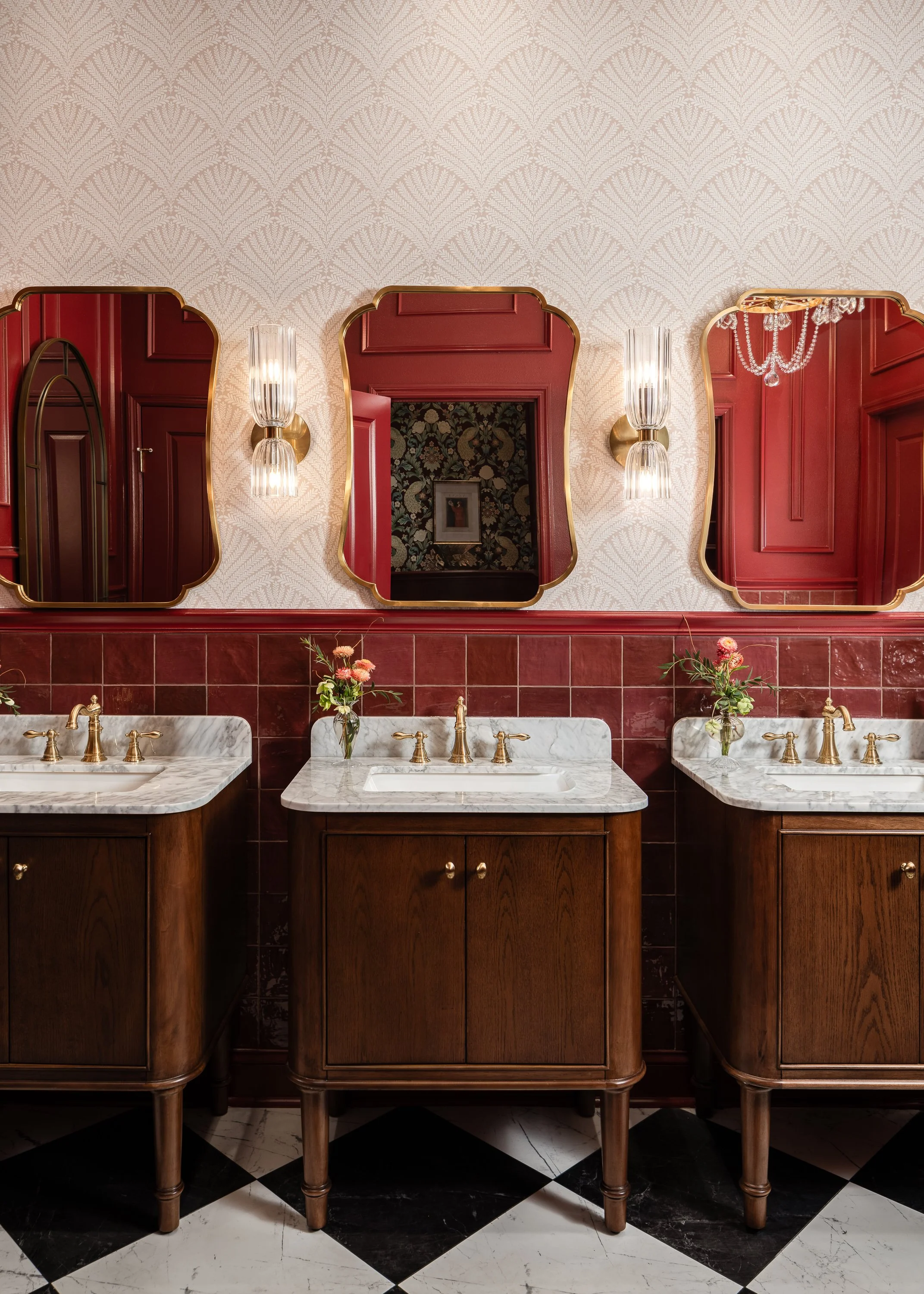 Three elegant vanity sinks in a commercial restroom deswith marble countertops and wooden cabinets are set against a wall with red tile and patterned wallpaper. Ornate gold-framed mirrors hang above each sink, reflecting the room's luxurious details.