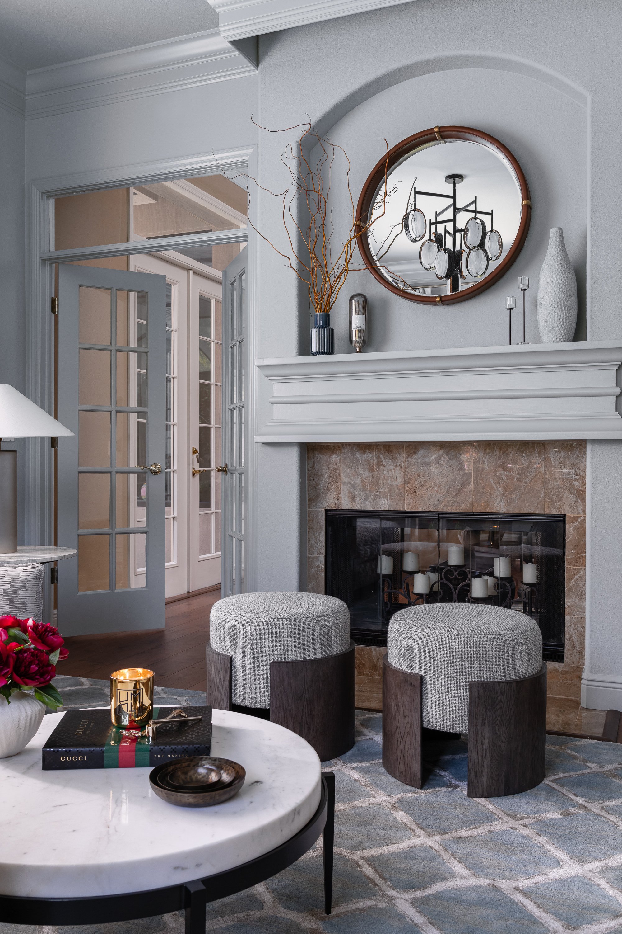 Contemporary modern sunroom design A living room features a fireplace with a round mirror above it, flanked by two grey upholstered stools. In the foreground, a marble coffee table holds books and a vase of red flowers.