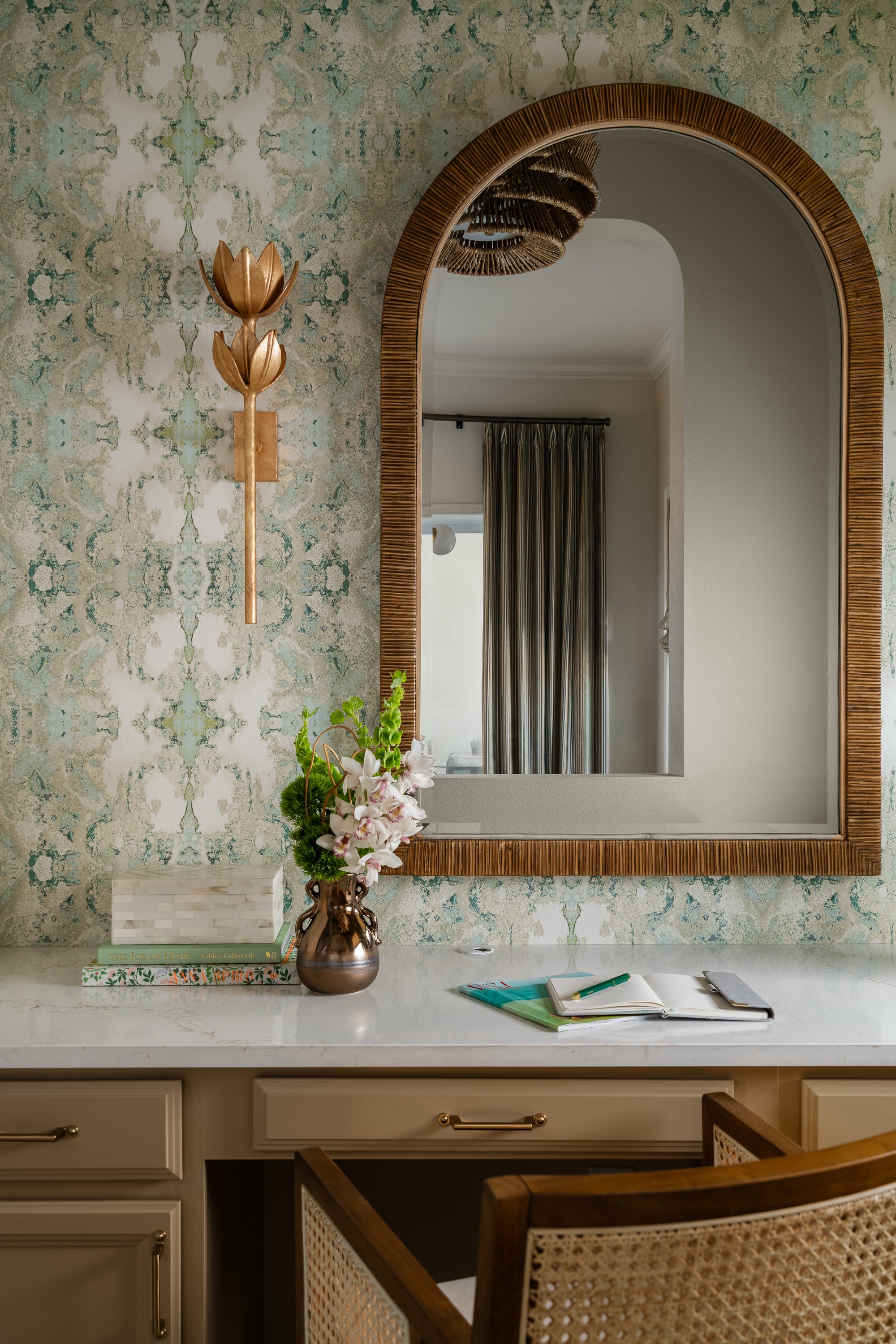 A vanity counter with a marble top, featuring a vase with flowers, stacked books, a notebook, and an arched mirror with a woven frame. A gold floral sconce is mounted on the patterned wallpaper above.
