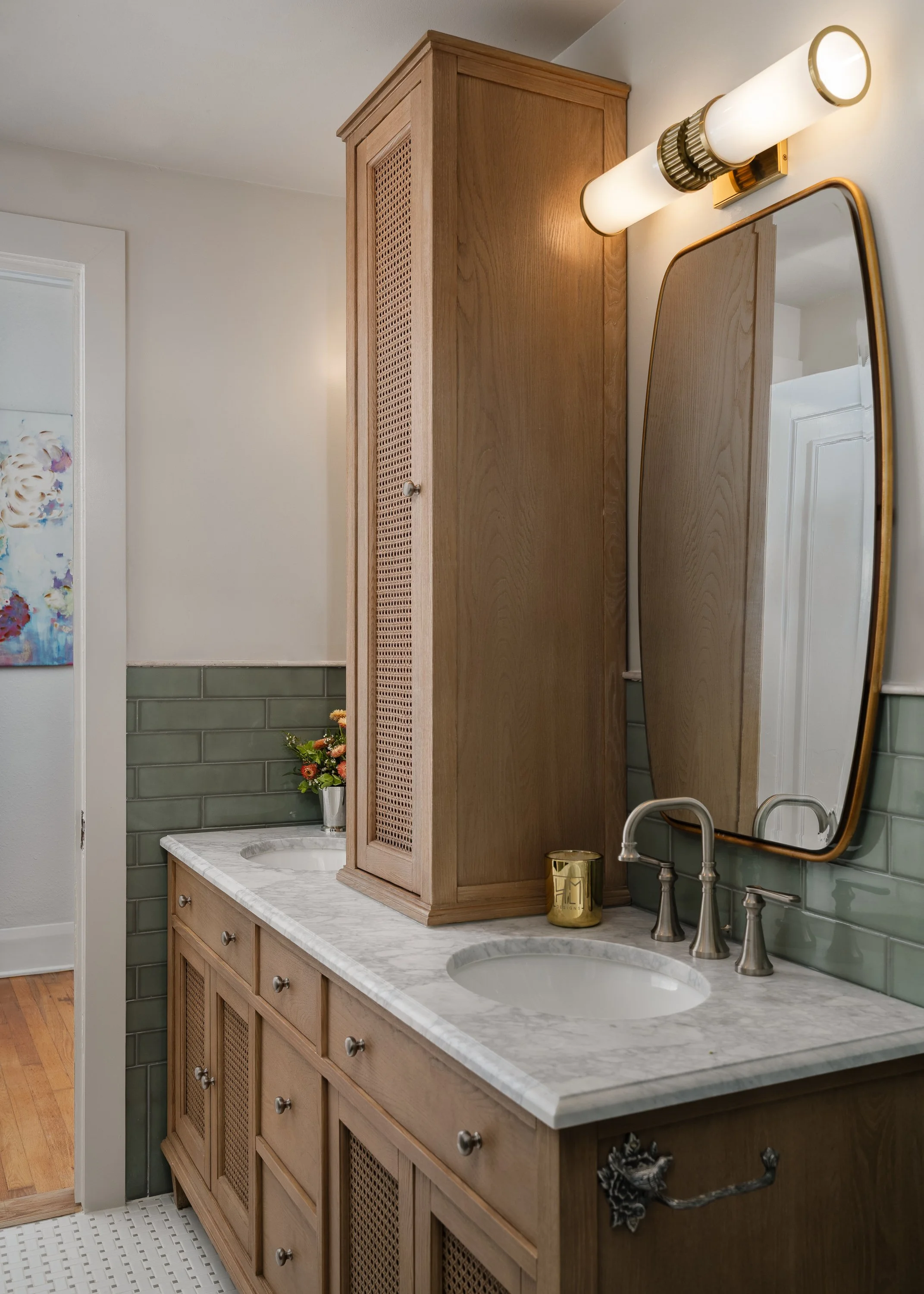 A modern bathroom vanity with a marble countertop, two sinks, and a tall wooden cabinet with cane detailing. A gold-framed mirror hangs above the vanity, and a cylindrical light fixture illuminates the space.