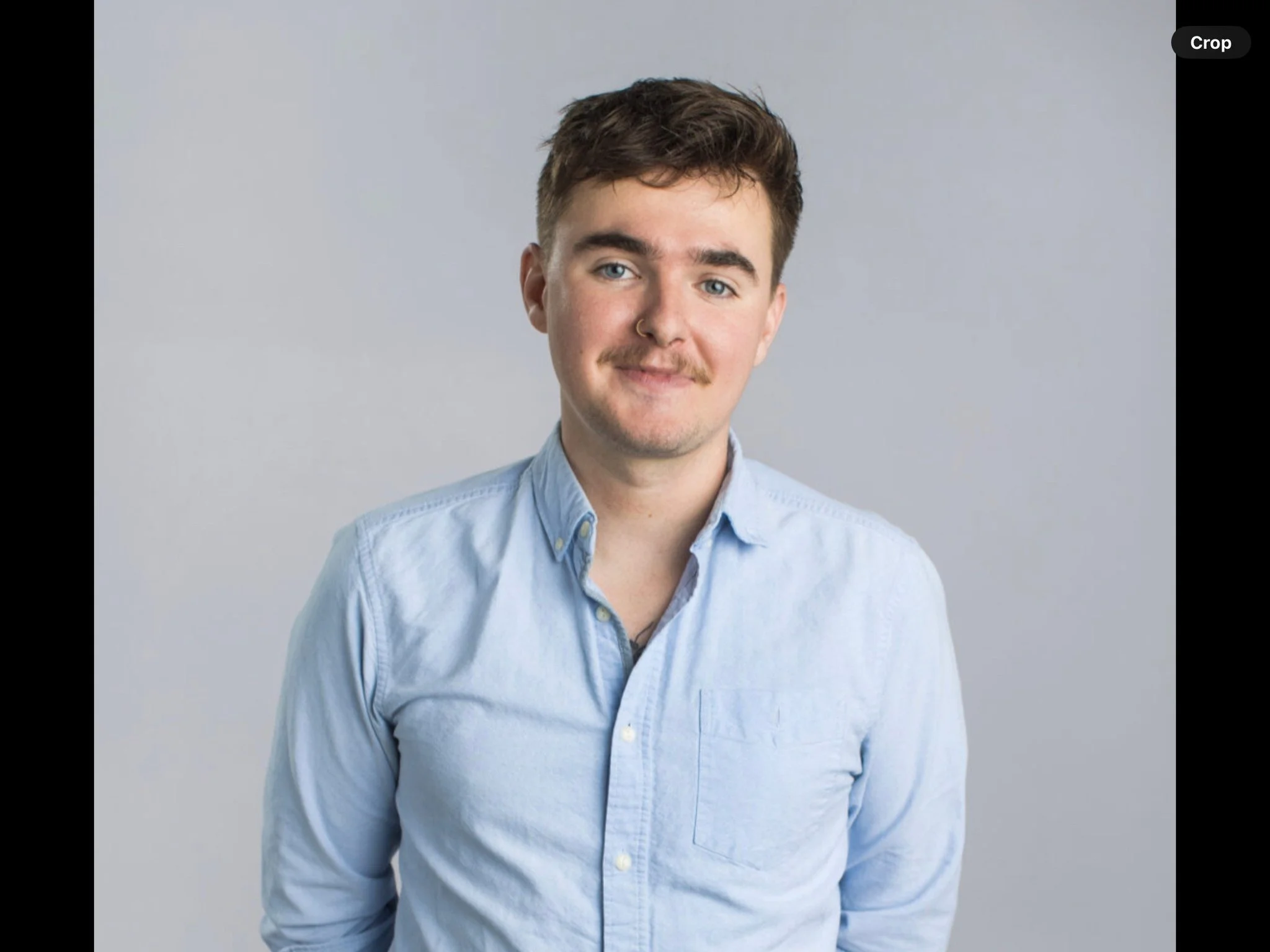A young man with brown hair, blue eyes, and a light mustache, wearing a light blue button-up shirt, standing against a plain gray background.