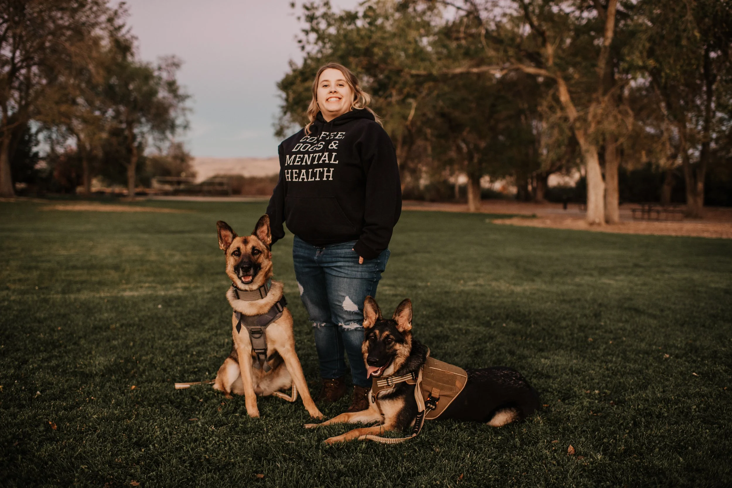 A woman smiling in a park with two dogs, one sitting and one lying down, both wearing harnesses, during sunset.