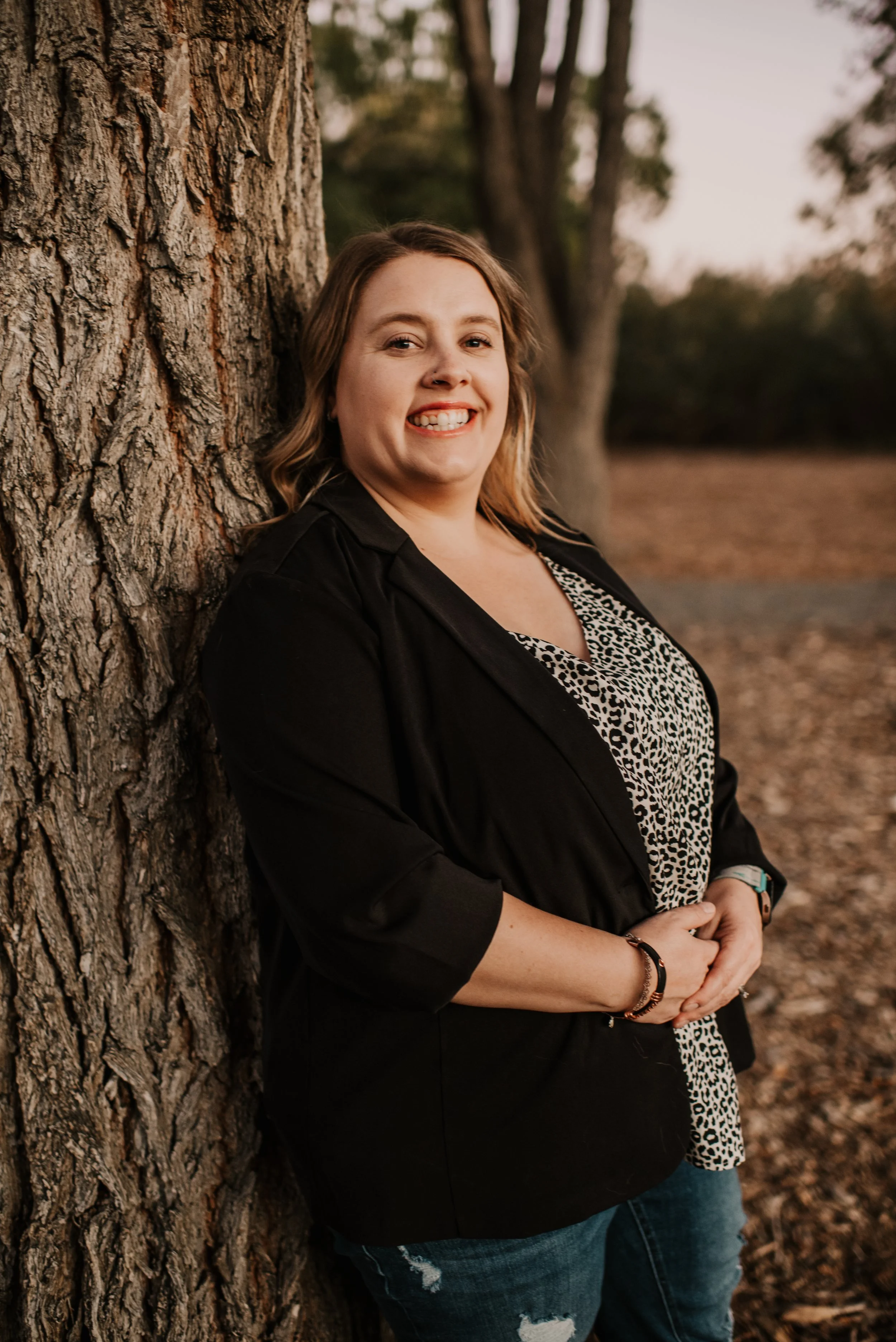 A woman standing outdoors leaning against a large tree with a textured bark, smiling, during golden hour.