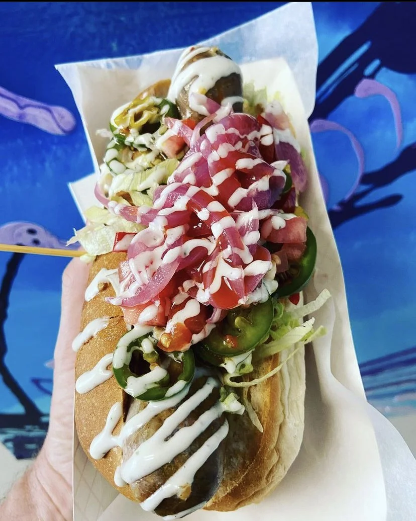A hot dog topped with lettuce, sliced jalapeños, pickles, chopped tomatoes, shredded lettuce, and drizzled with mayonnaise and ketchup, served in a bun.
