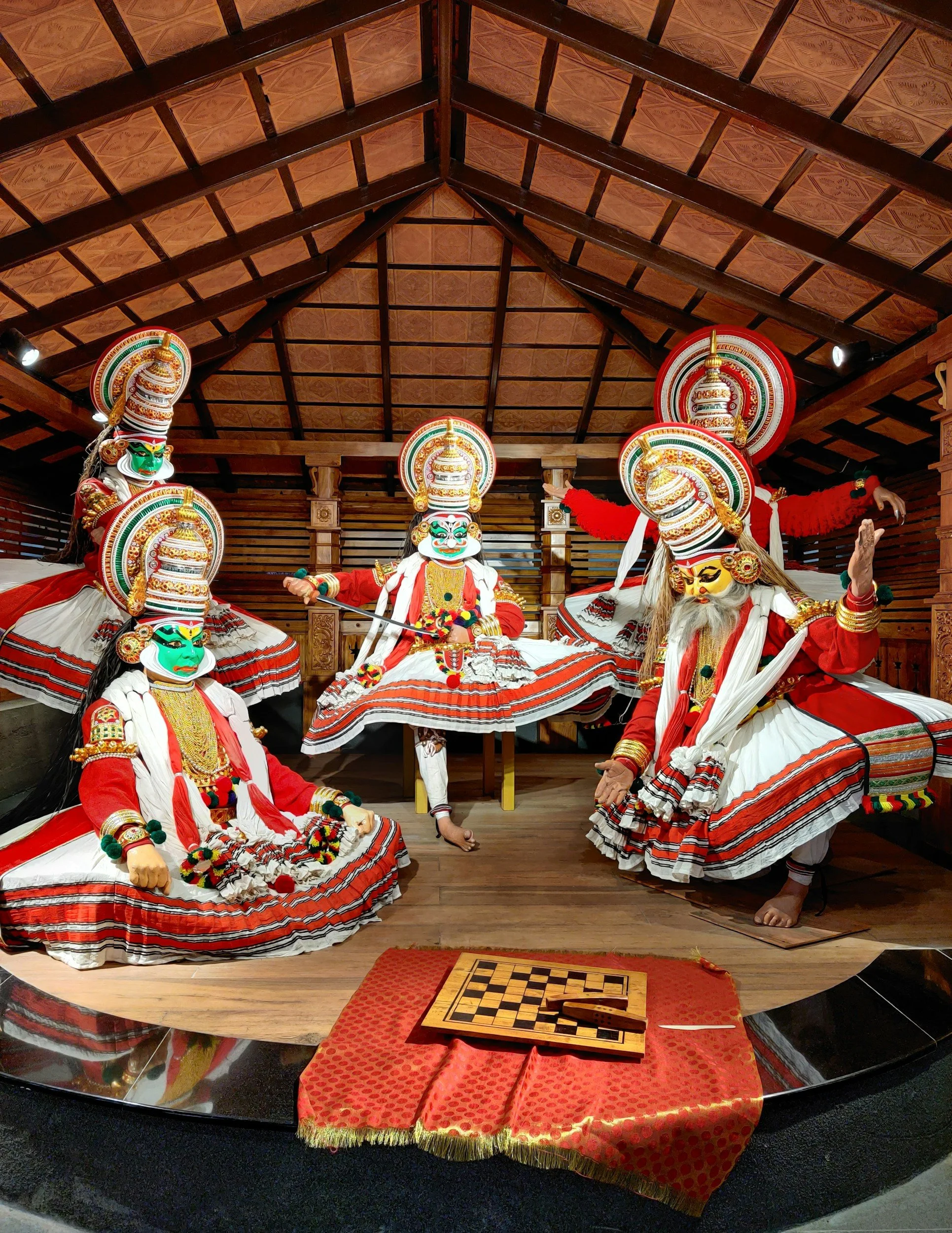 Colorful Kathakali performers in traditional costumes and masks in a wooden indoor setting with a chessboard on the floor.