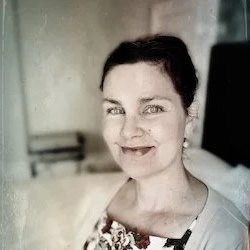 A woman with short dark hair smiling, wearing a floral top, in an indoor setting with a blurred background.