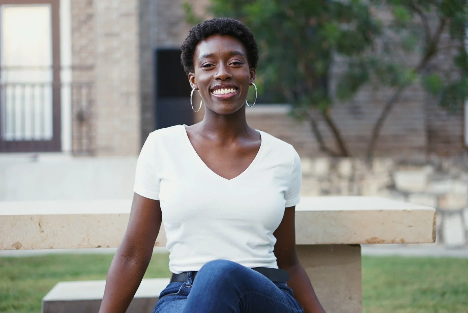 A young woman wearing a white V-neck T-shirt and hoop earrings, sitting outdoors on a concrete bench with a smile.