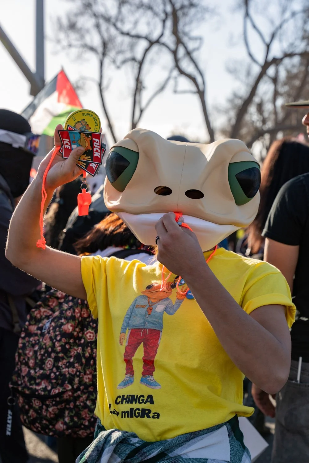 Person wearing a frog-shaped mask and a yellow t-shirt with a cartoon character on it, holding a whistle and protest badge, amid a crowd at an outdoor event.
