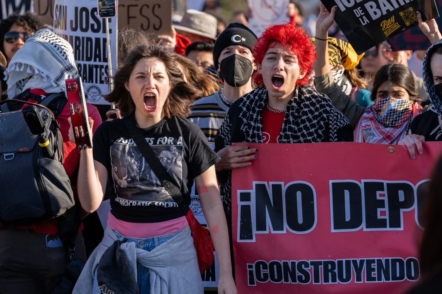 Group of people protesting, with two centered passionately shouting, one with red hair and the other with brown hair, holding a red banner  amid a crowd holding signs and wearing masks.