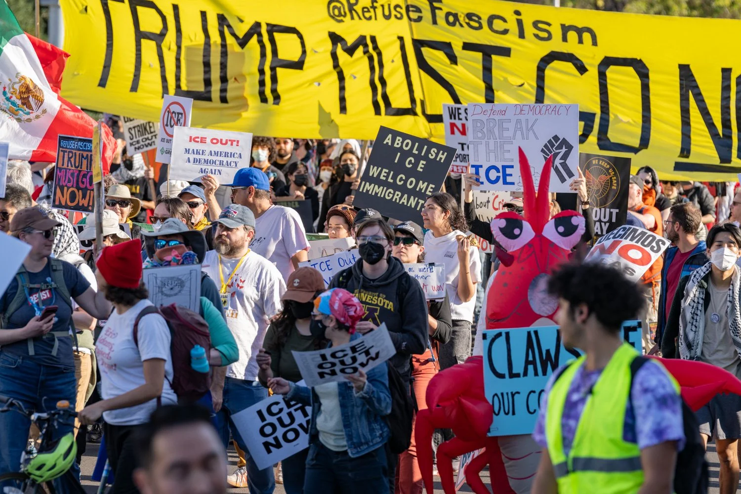 A large crowd of protesters at a rally holds various signs and banners, including a yellow banner at the top that says '@Refuse Fascism TRUMP MUST GO NOW,' and signs with messages like 'ICE OUT! MAKE AMERICA A DEMOCRACY AGAIN,' 'ABOLISH ICE,' 'BREAK 