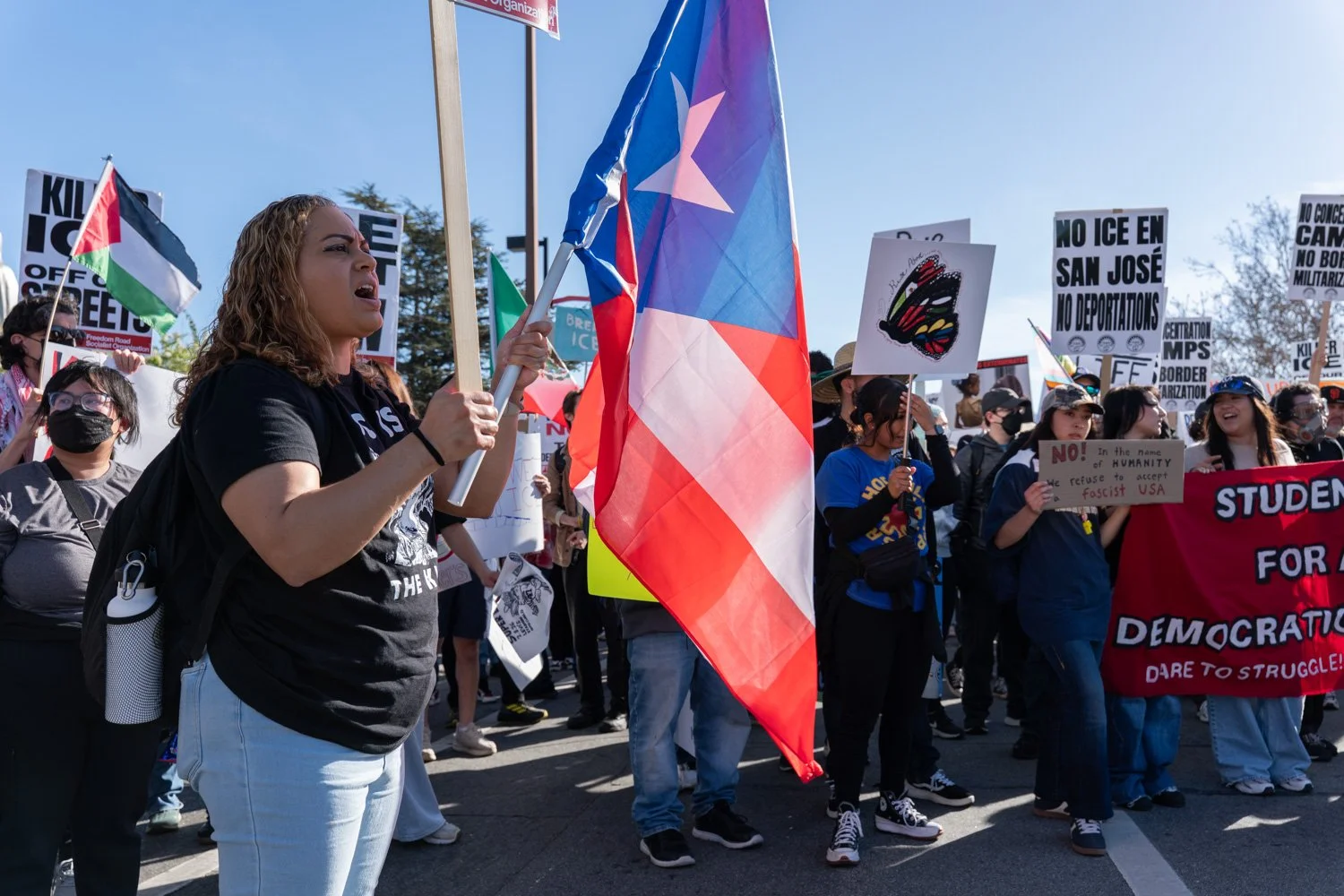 A woman with curly hair holding a flag and shouting at a protest rally. She is surrounded by other protesters holding signs and flags, some wearing masks, advocating against deportations and supporting human rights.