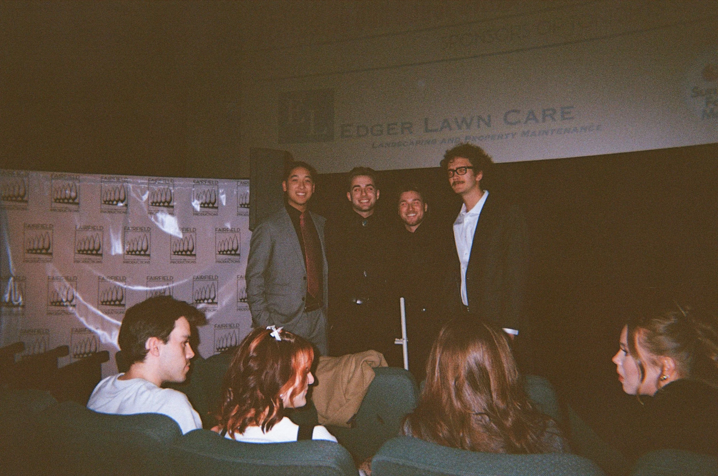 A group of five men standing on a stage with a black backdrop, while an audience watches. The stage is at Fairfield Productions, and there is a sign for Edger Lawn Care in the background.
