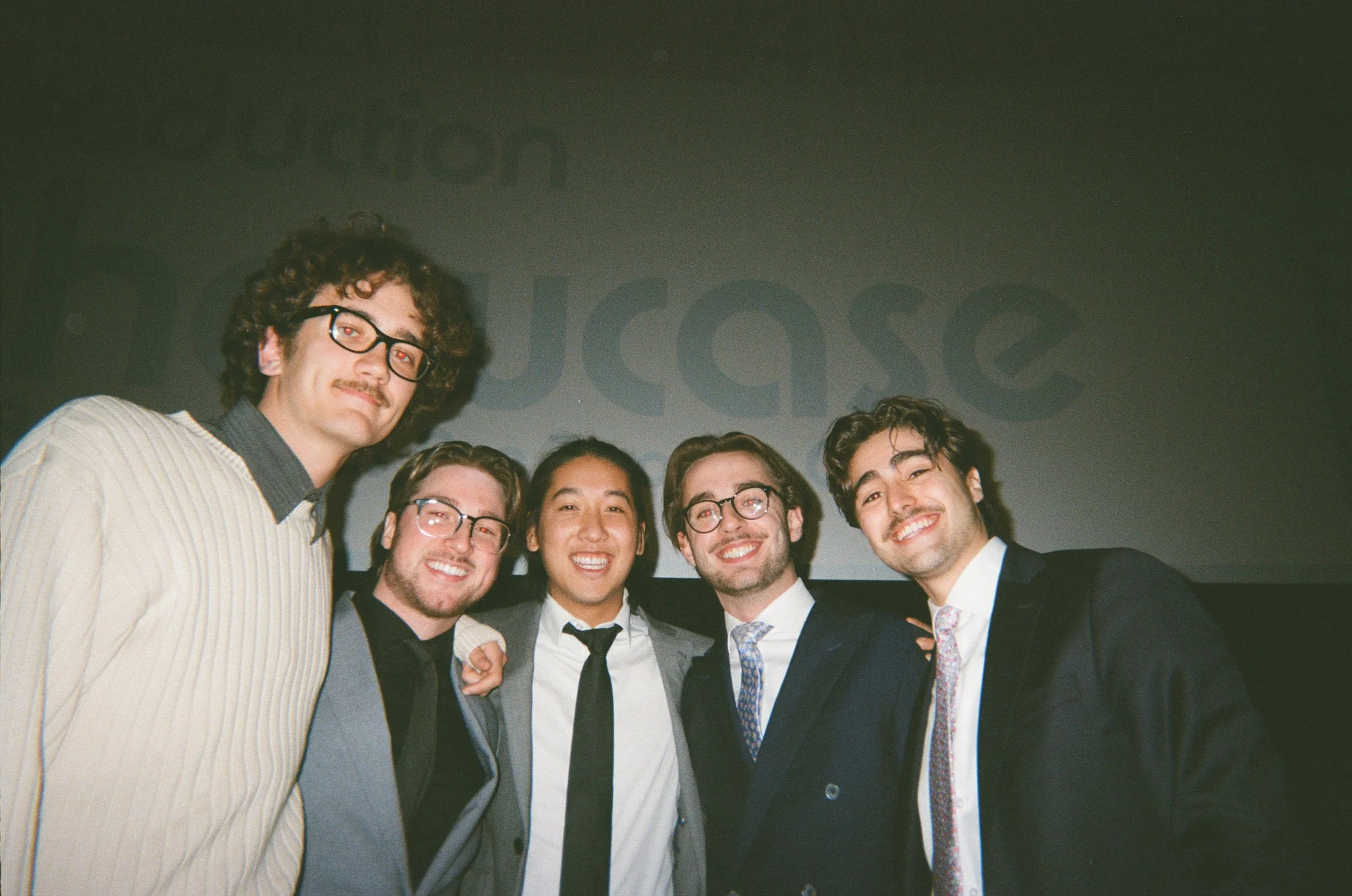 Group of five young men in formal attire smiling at the camera in front of a dark background with a faint projection that reads 'Juse'.
