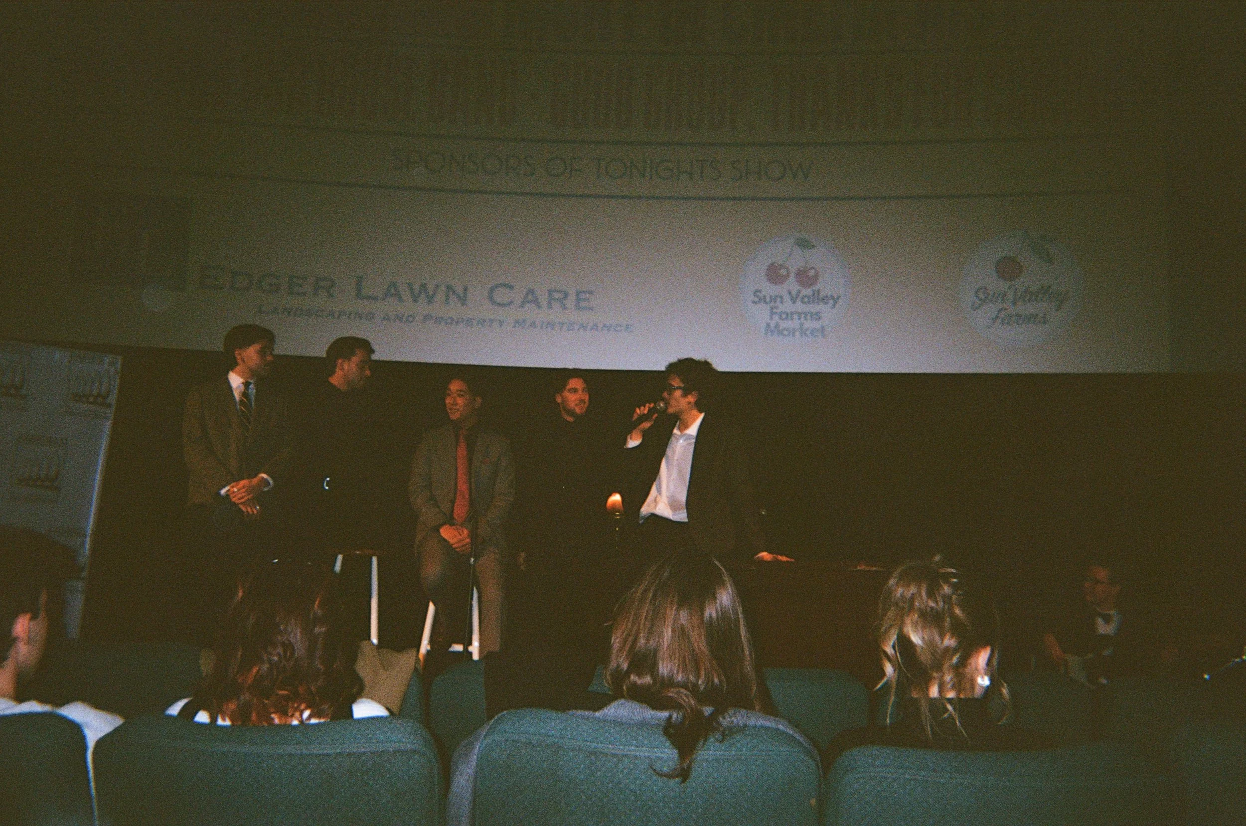 A panel of five men in formal attire on stage during a presentation, with an audience seated in front of them. A large screen behind the panel displays logos and text.
