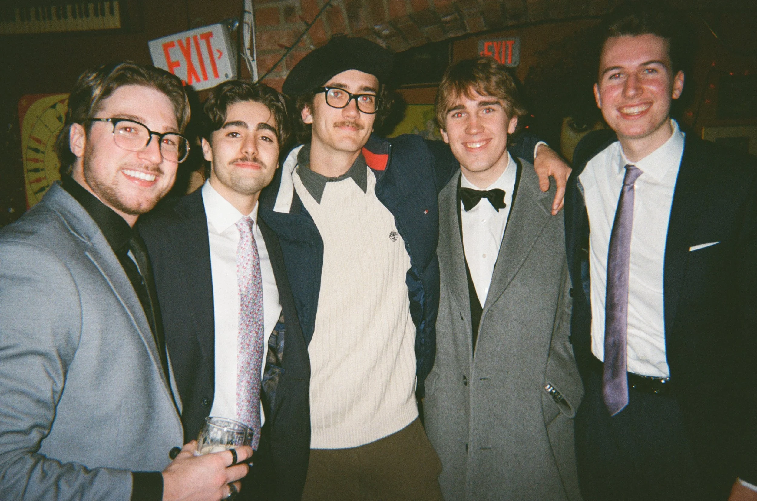 Group of five men in formal and semi-formal attire posing together at an indoor event, with exposed brick walls and illuminated exit signs in the background.