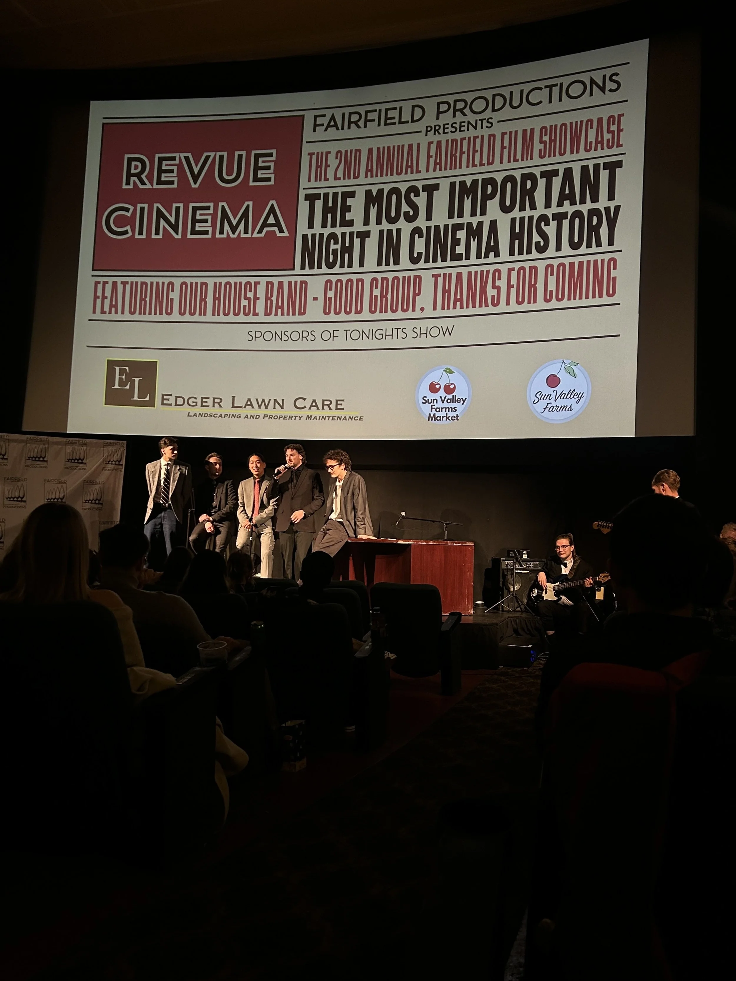 A group of performers on stage at a film showcase event, with a large screen behind them displaying event details and logos, including sponsors.