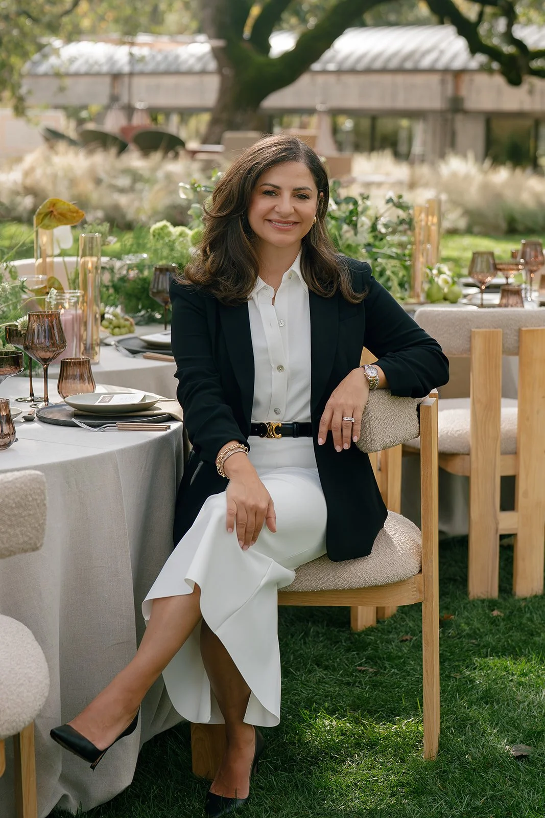 A woman sitting outdoors at a table decorated for a gathering, wearing a black blazer over a white shirt and white pants, smiling at the camera.