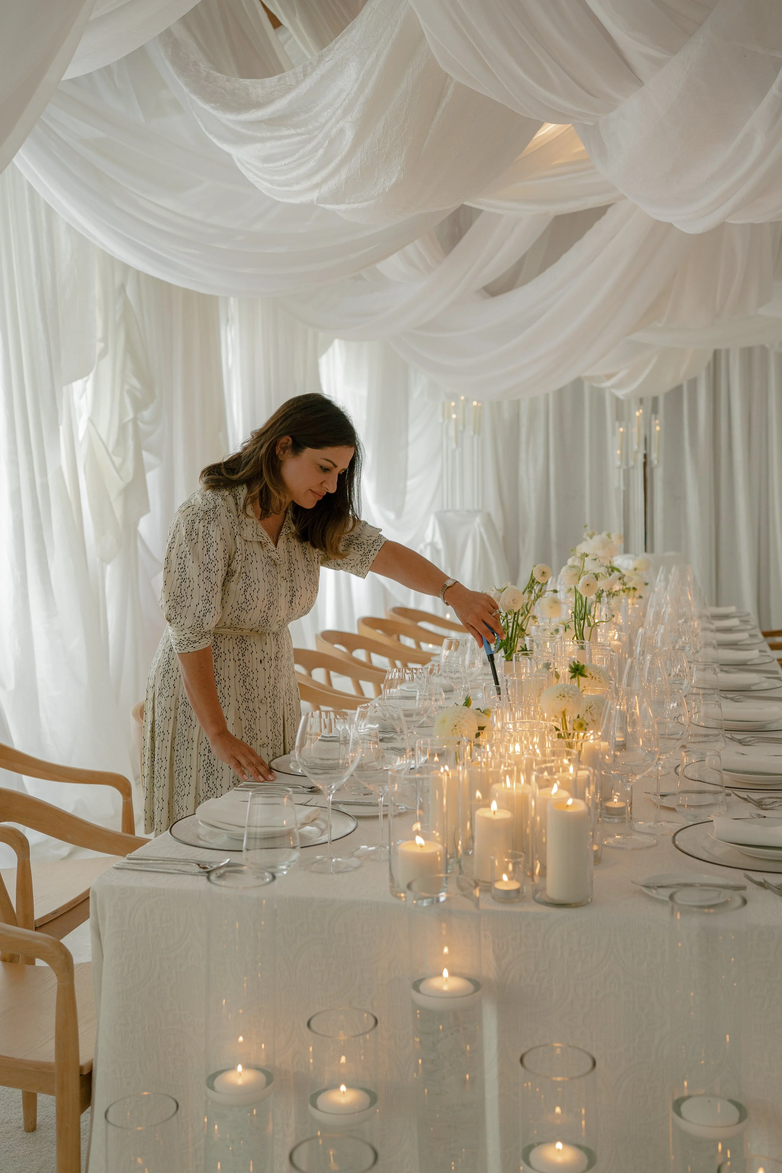 A woman arranging flowers on a decorated banquet table in a white-draped elegant venue with candles and glassware.