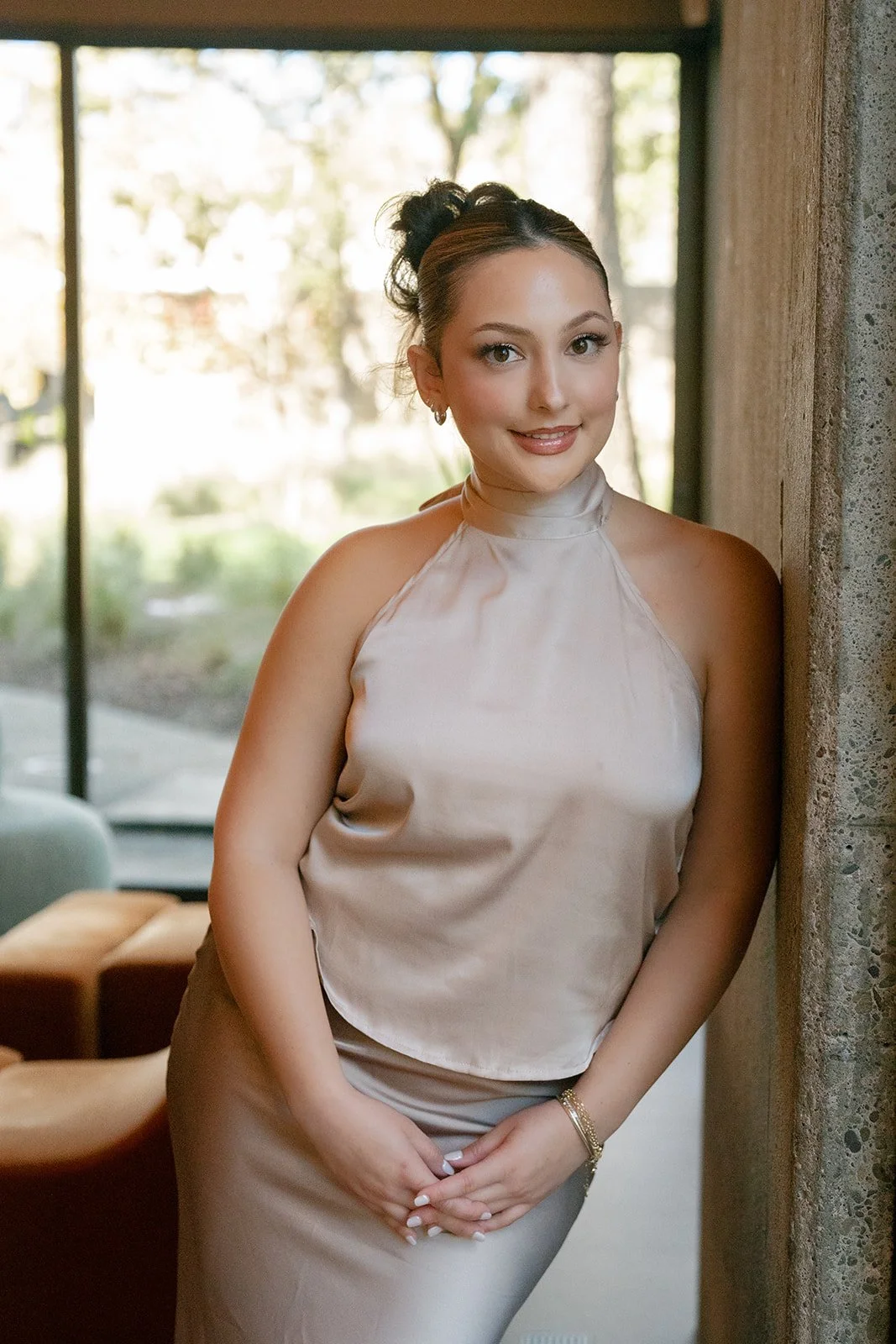 A young woman with curly hair styled in an updo, wearing a satin sleeveless top and matching skirt, stands indoors near a concrete wall with a large window behind her revealing an outdoor scene, smiling softly at the camera.