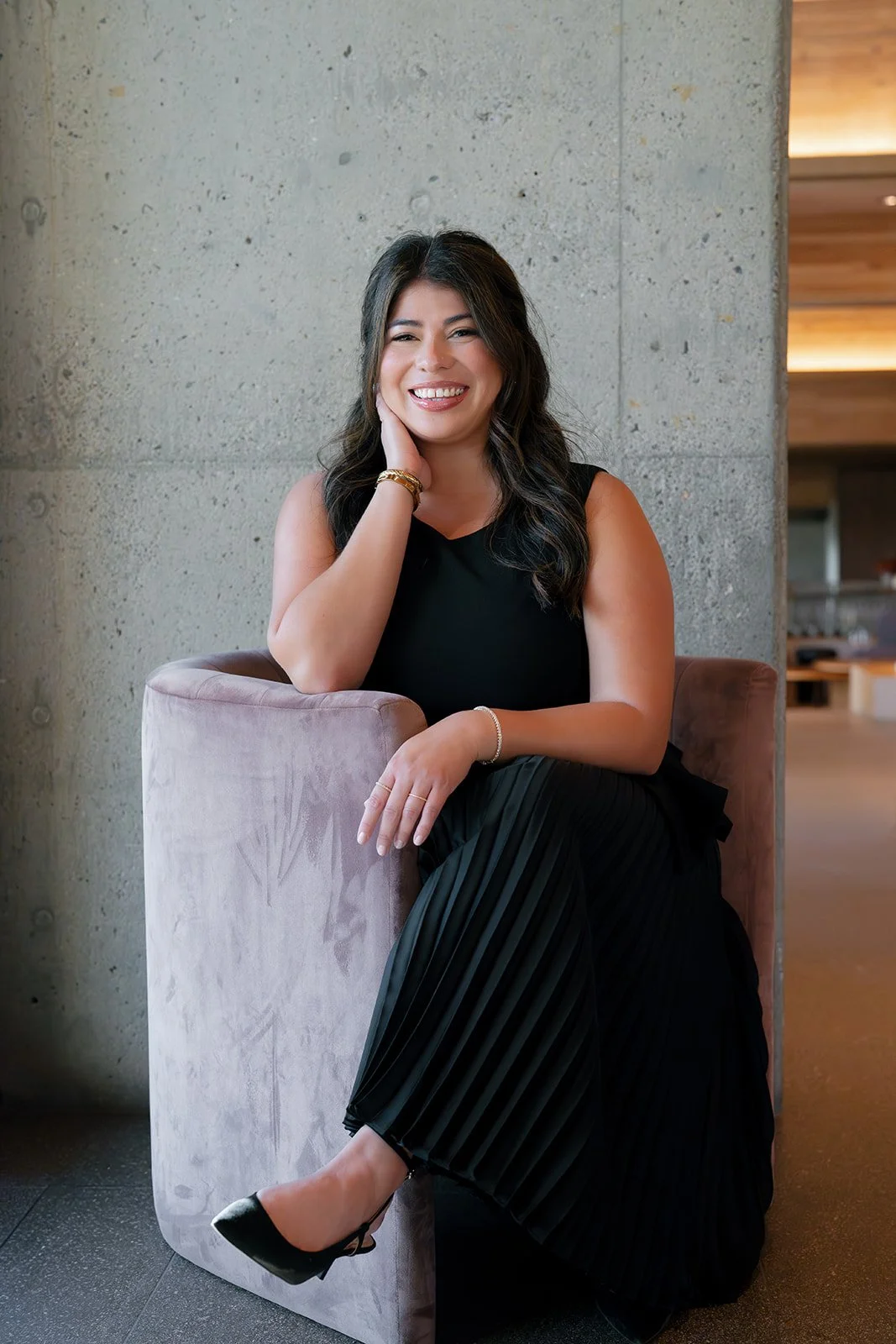 A woman with long dark hair smiling while sitting on a pink velvet armchair against a concrete wall, wearing a sleeveless black top, black pleated skirt, and black heels, in a modern interior setting.