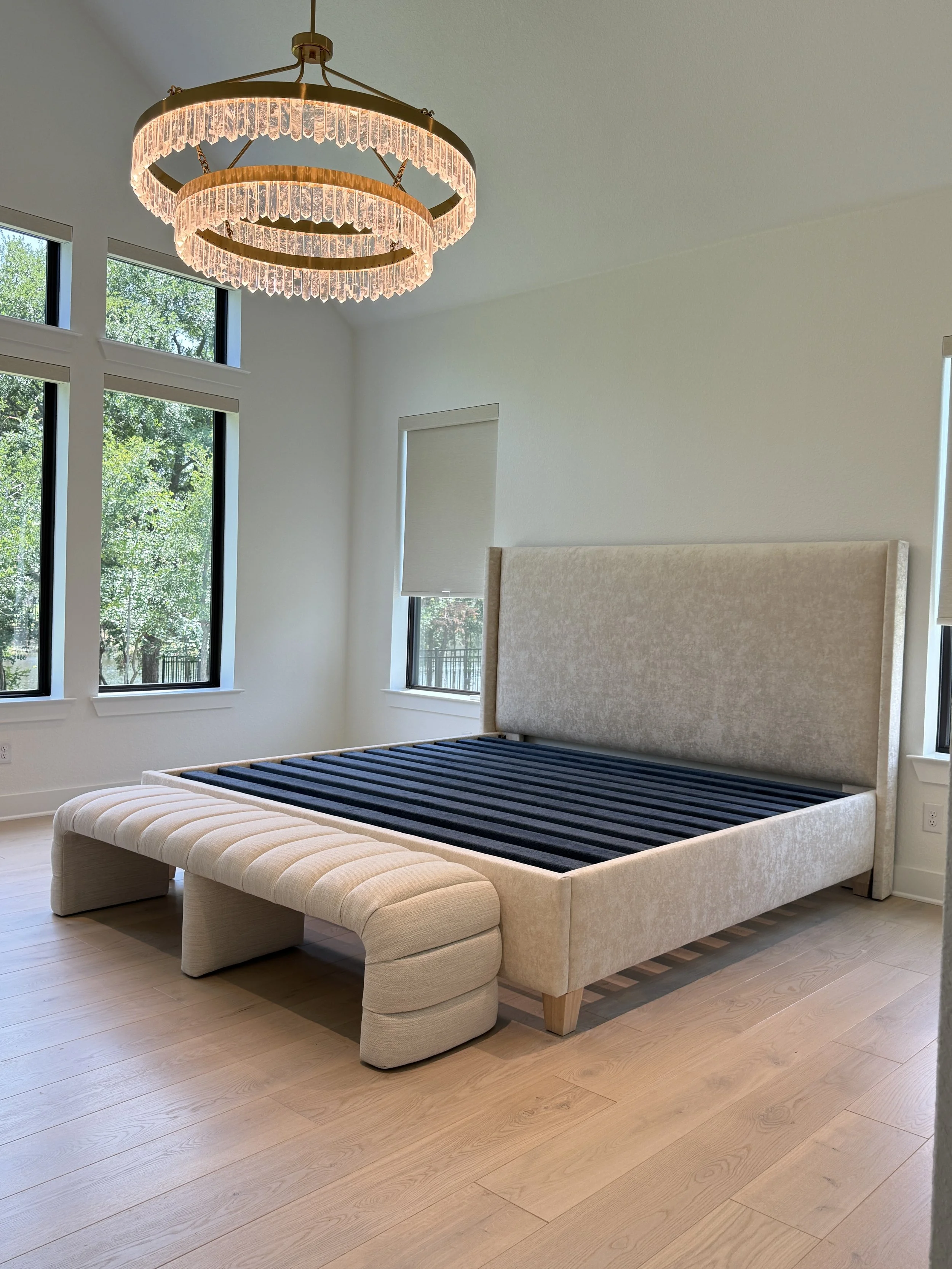 An empty beige upholstered bed frame with a matching bench at the foot, set in a bright room with wooden floors, large windows, and a modern circular chandelier with crystal accents hanging from the ceiling.