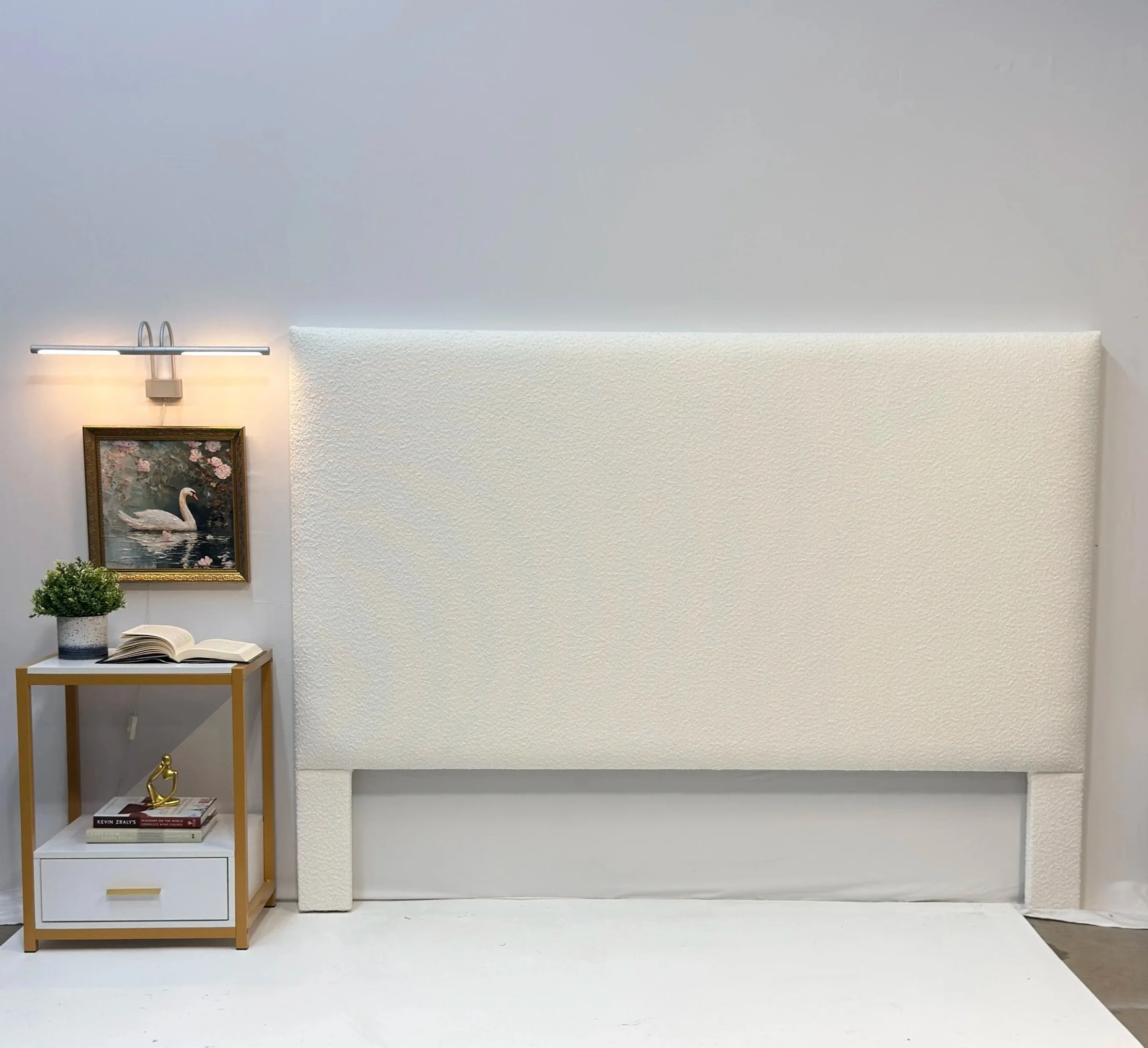 Minimalist and modern room with a white textured fireplace, a wooden bookshelf with books, a small potted plant, a framed painting of a swan, a gold sculpture, and a wall-mounted light fixture.