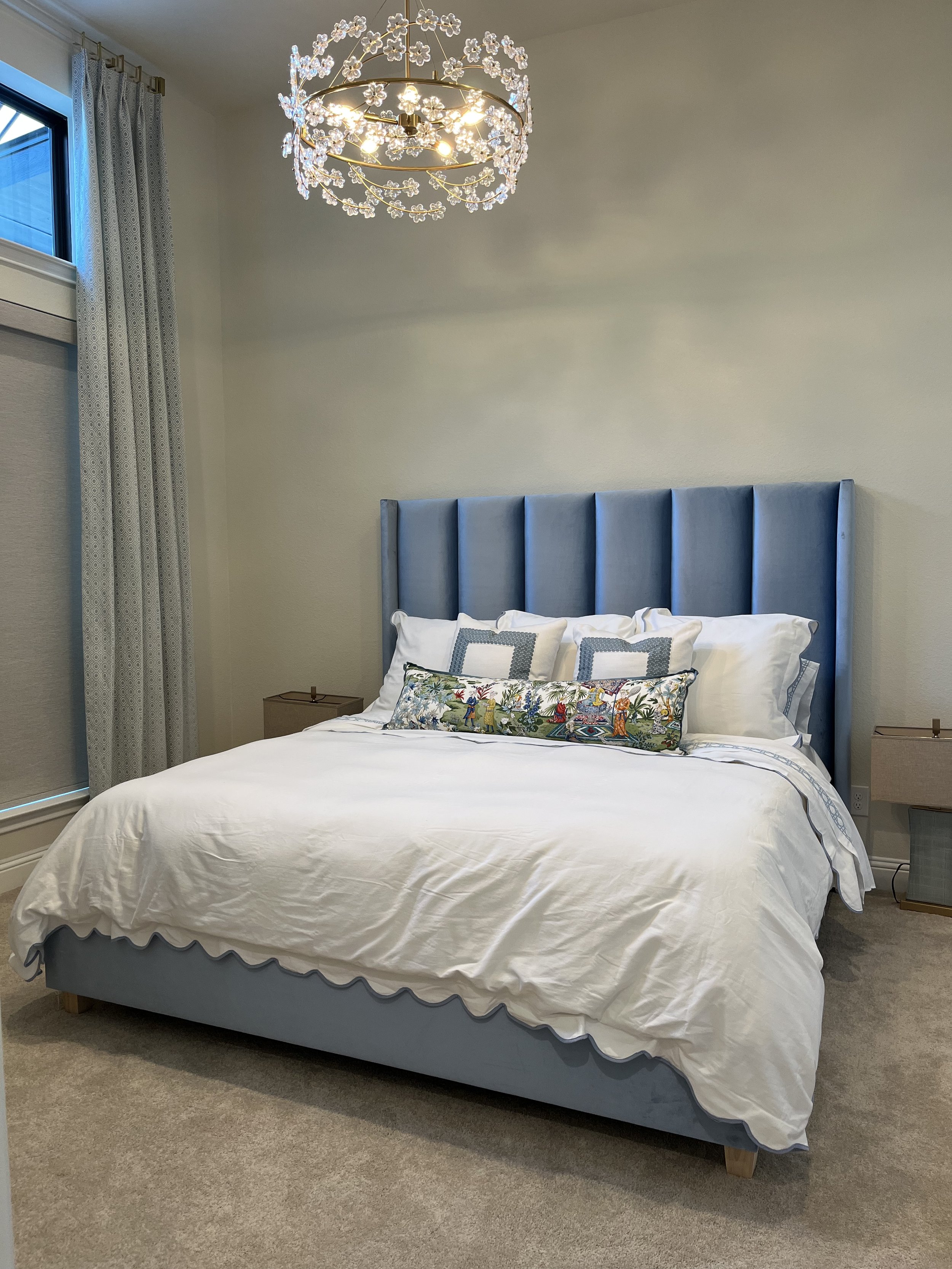A bedroom featuring wingback design tufted blue upholstered headboard, white bedding, and decorative pillows. There are two nightstands, a window with light-colored curtains, and a floral-patterned pillow on the bed.