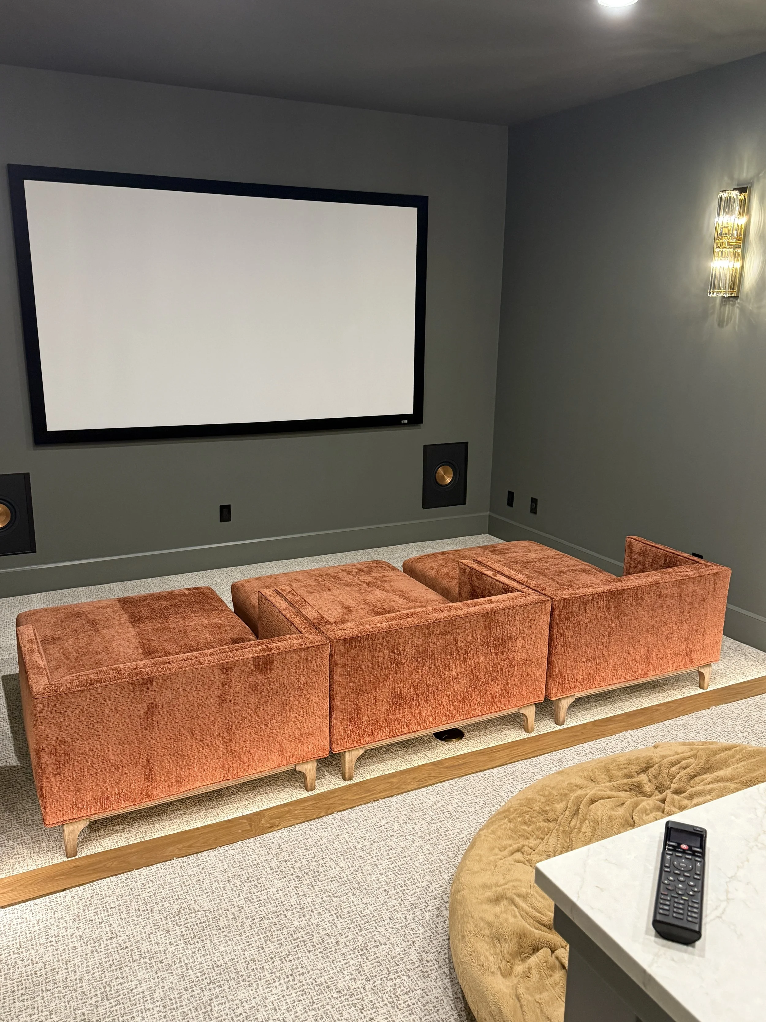 A cozy home theater room with a beautiful reupholstered, peach-colored velvet chaise lounges, a remote control on a white marble table, and soft lighting.
