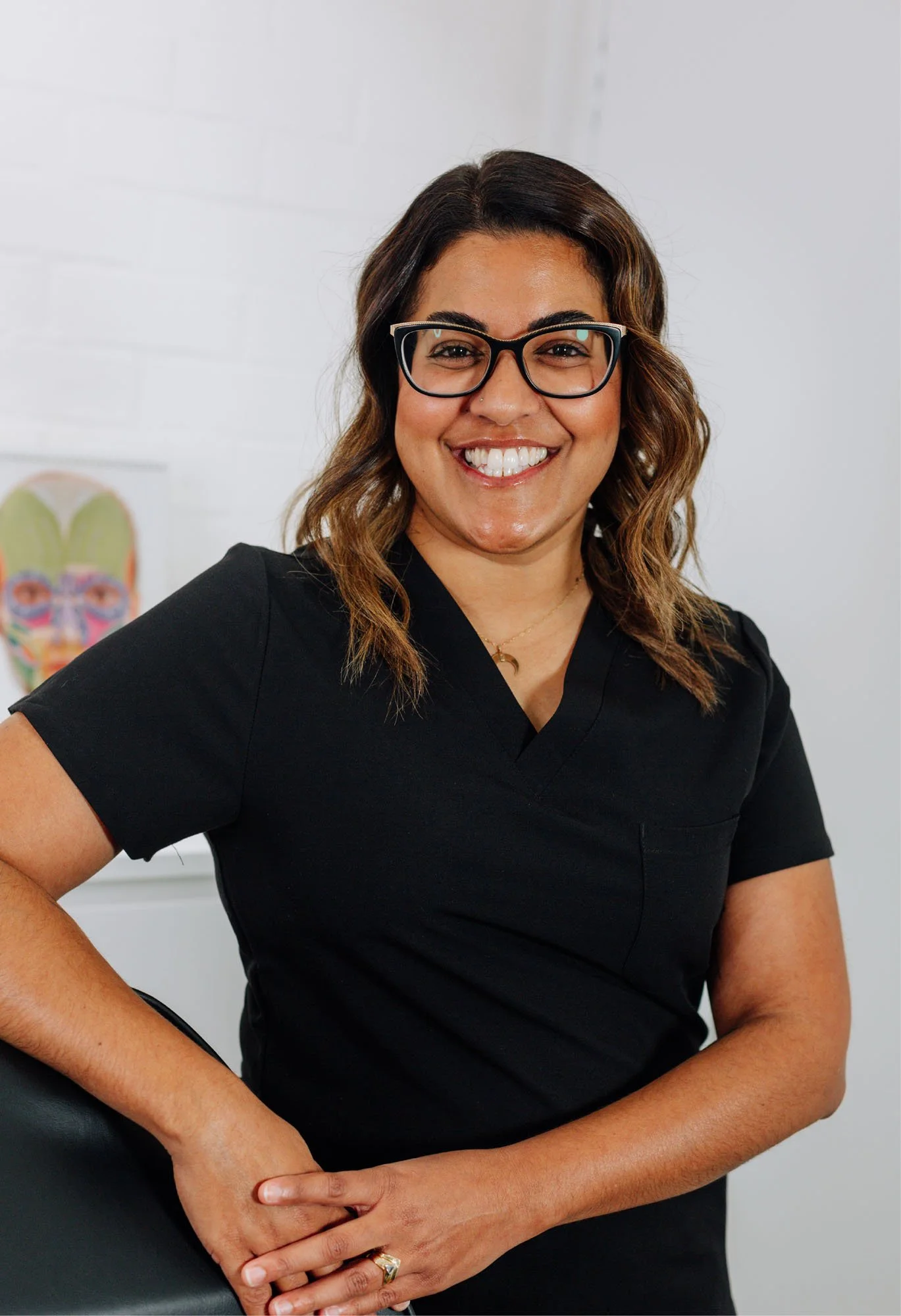 Close-up of a smiling woman with glasses, wearing a black top, seated at a desk with a human head anatomical illustration in the background.
