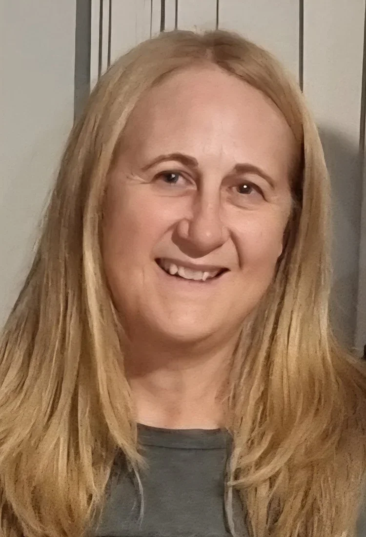 A woman with long blonde hair, smiling, wearing a gray shirt, with a wooden headboard in the background.