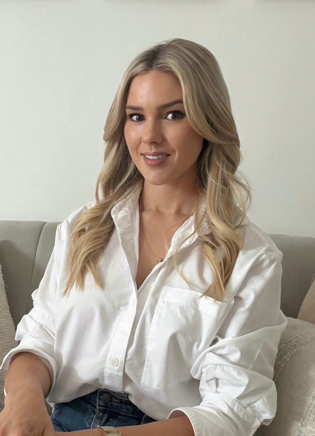 A young woman with blonde hair styled in loose waves, wearing a white button-up shirt, smiling, sitting on a light-colored sofa with pillows, against a plain white wall.