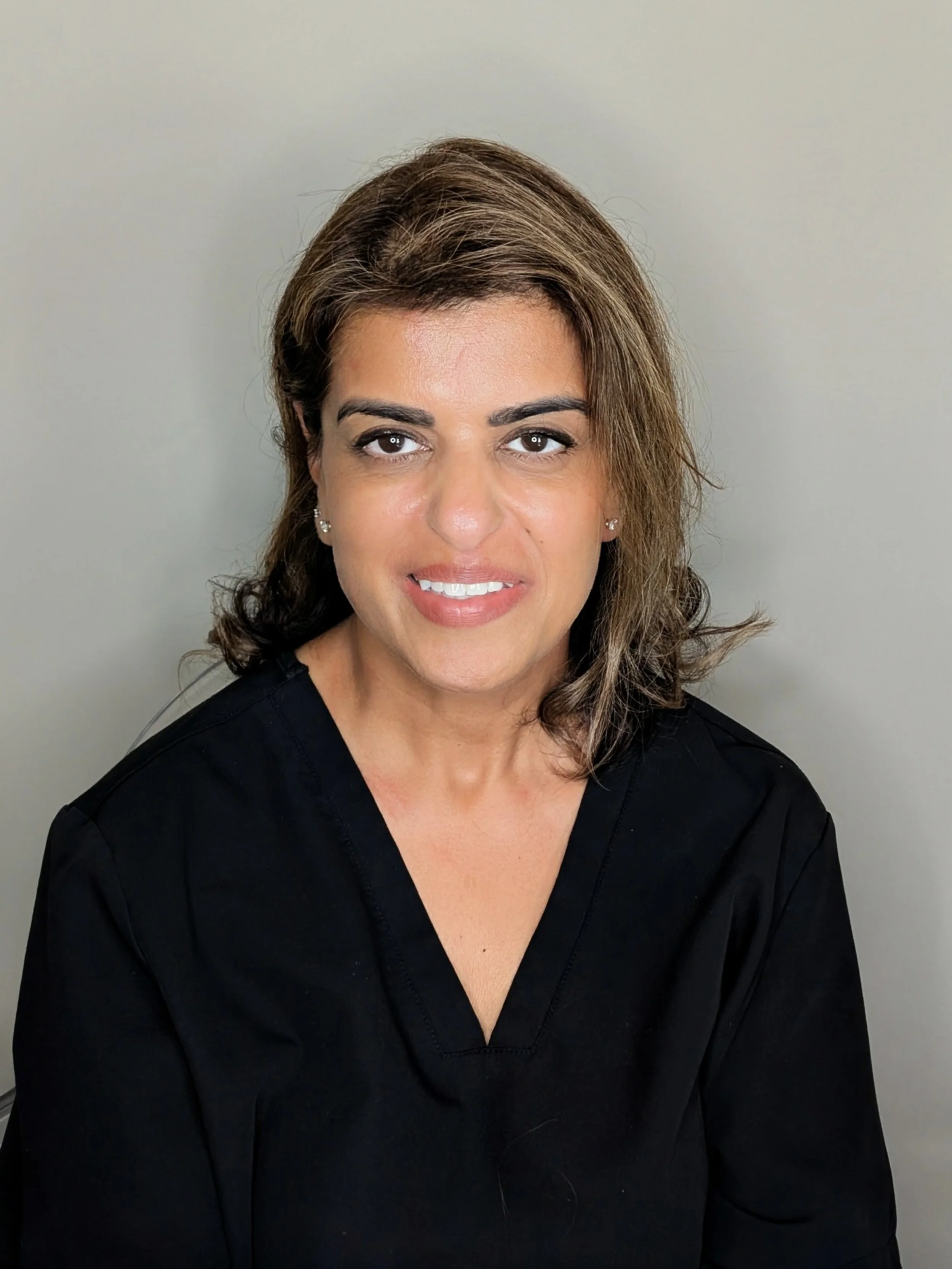 A woman wearing a black medical scrub top, with shoulder-length brown hair with lighter highlights, smiling in front of a plain light-colored wall.