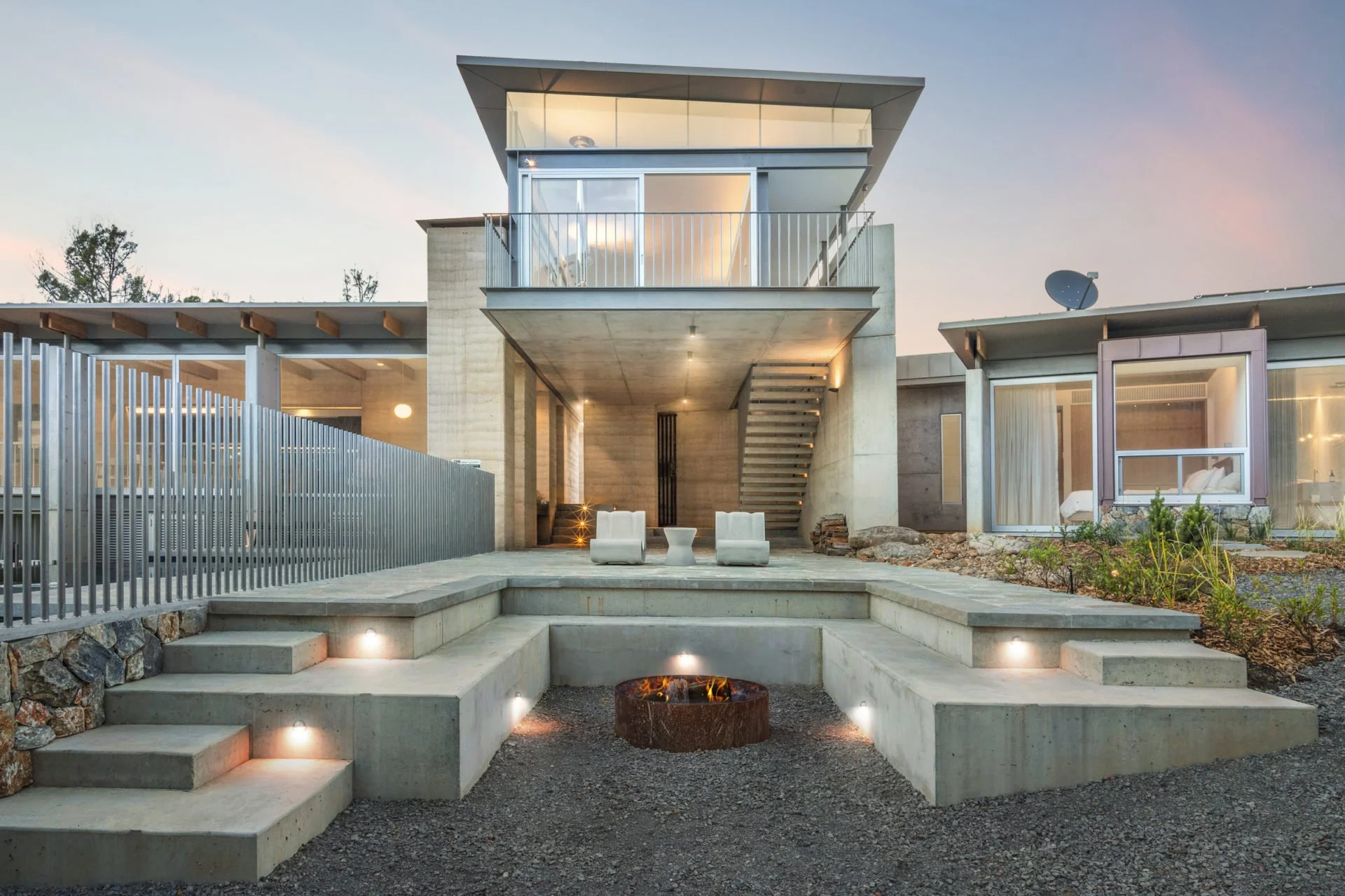 Modern house exterior with concrete and glass design, featuring a multi-level outdoor seating area with steps and integrated lighting, a central fire pit, and an external staircase leading to an upper balcony.
