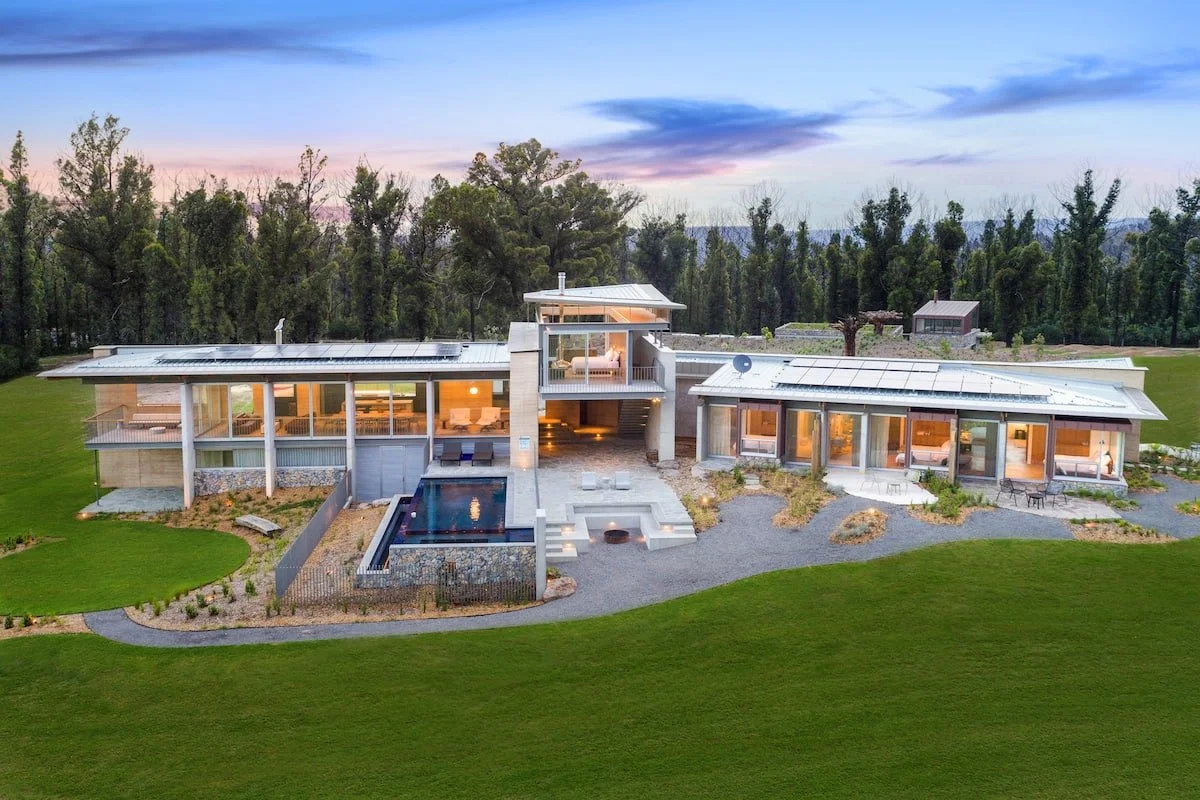 Modern luxury house with large windows, a central courtyard, swimming pool, and landscaped garden, surrounded by trees, under a blue sky.