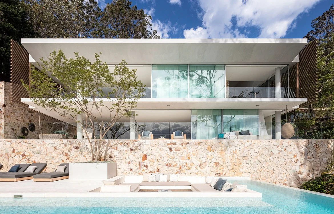 Modern two-story house with glass walls, surrounded by trees, featuring a stone facade, outdoor seating area, and swimming pool.