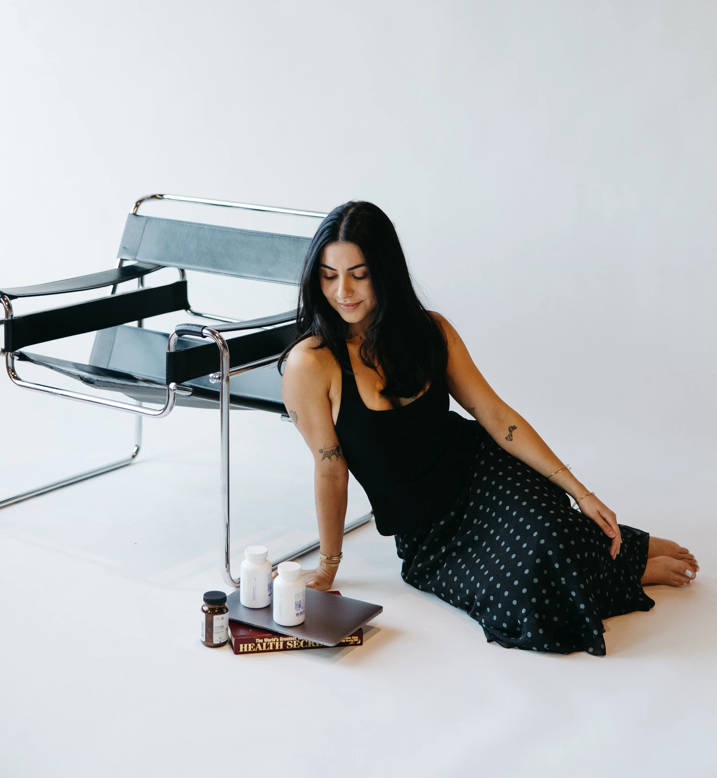Woman with dark hair sitting on the floor next to a metal chair, with medicine bottles, a book titled 'Health Secrets,' and a tablet in front of her.