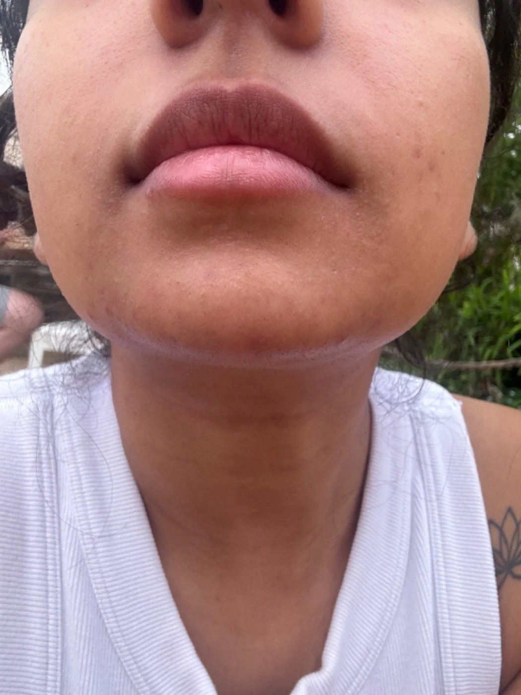 Close-up of a person's face showing lips, chin, and part of their nose, with a tattoo on one shoulder, wearing a white sleeveless top, outdoors with greenery in the background.