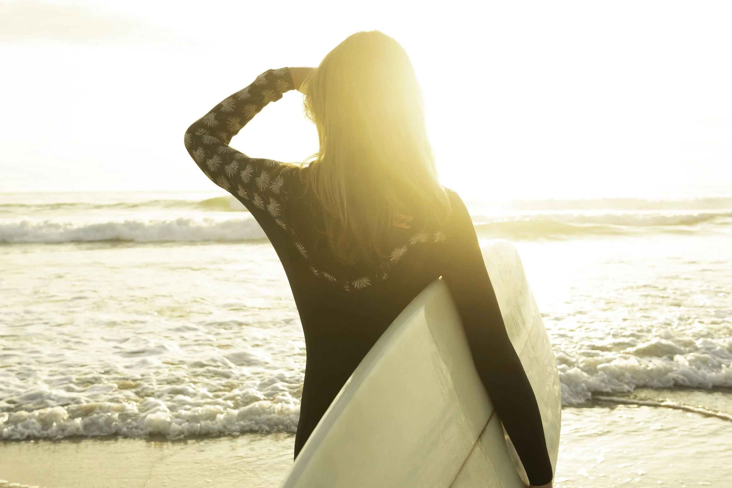 women looking out towards the ocean holding surfboard and shielding her eyes from the sun