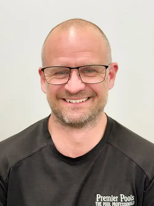 A smiling man with glasses and a short beard, wearing a black shirt that reads 'Premier Pools, The Pool Professionals,' standing against a plain light background.