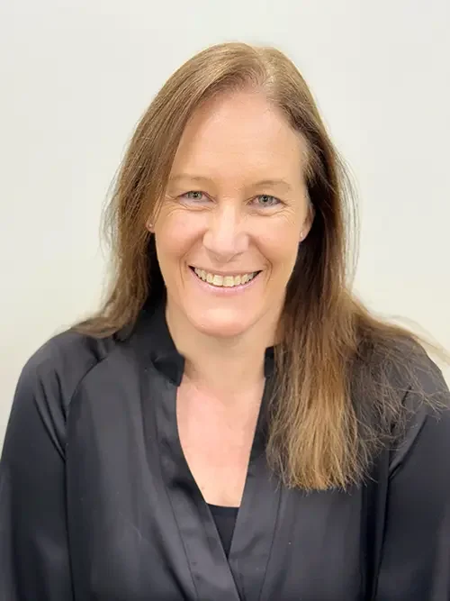 A woman with long, light brown hair smiling at the camera, wearing a dark gray or black blouse, standing against a plain light-colored background.