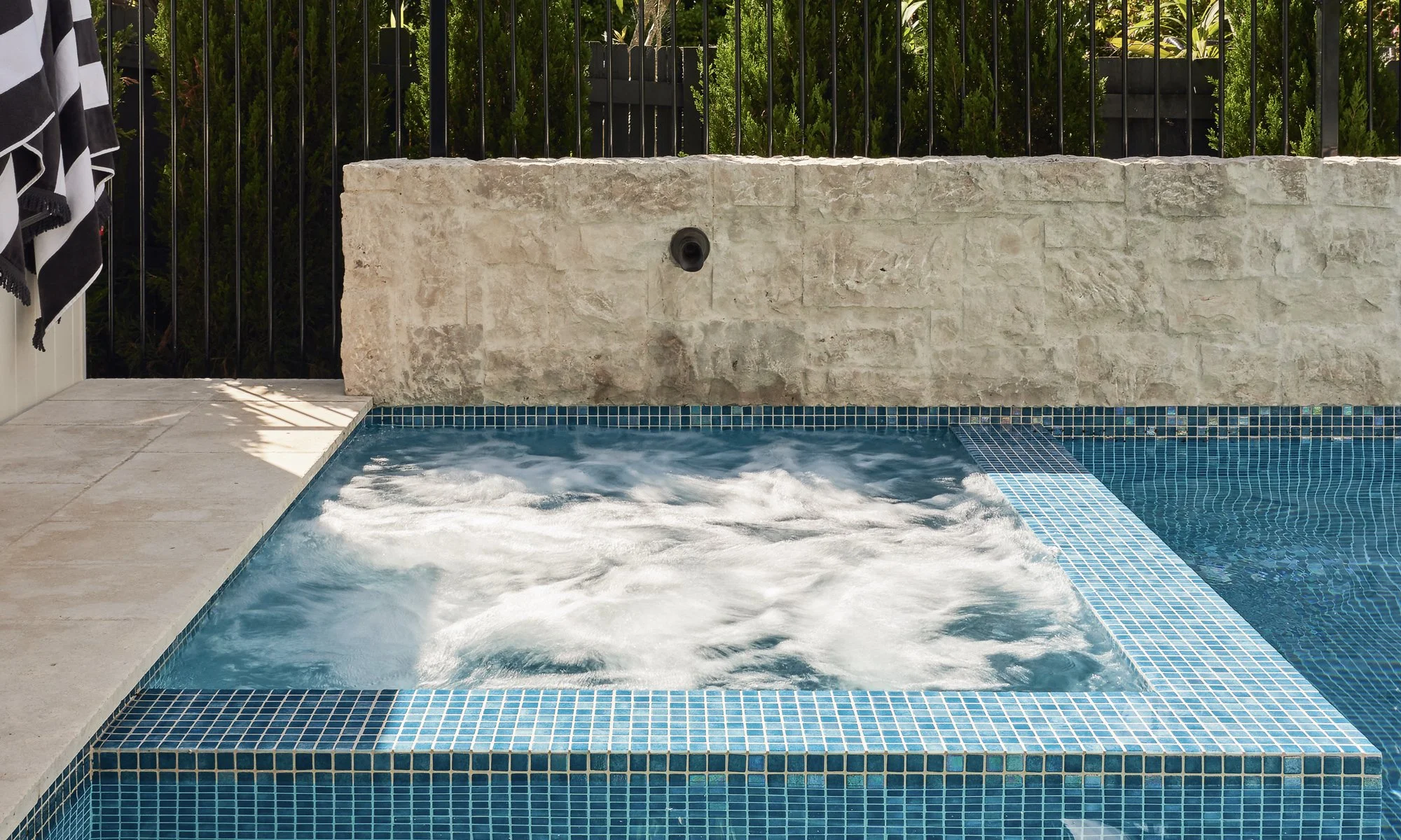 Close-up of a bubbling hot tub with blue mosaic tiles, adjacent to a swimming pool, with a stone wall and fencing in the background.