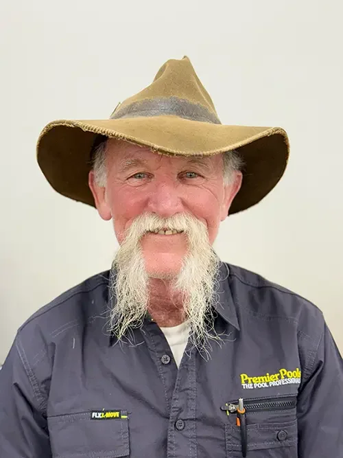 An older man with a long white beard and mustache smiling, wearing a wide-brimmed hat and a navy blue work shirt with the logo of Premier Pools.