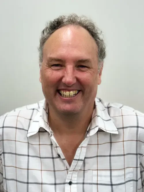 A smiling man with curly hair wearing a white plaid shirt against a plain off-white background.