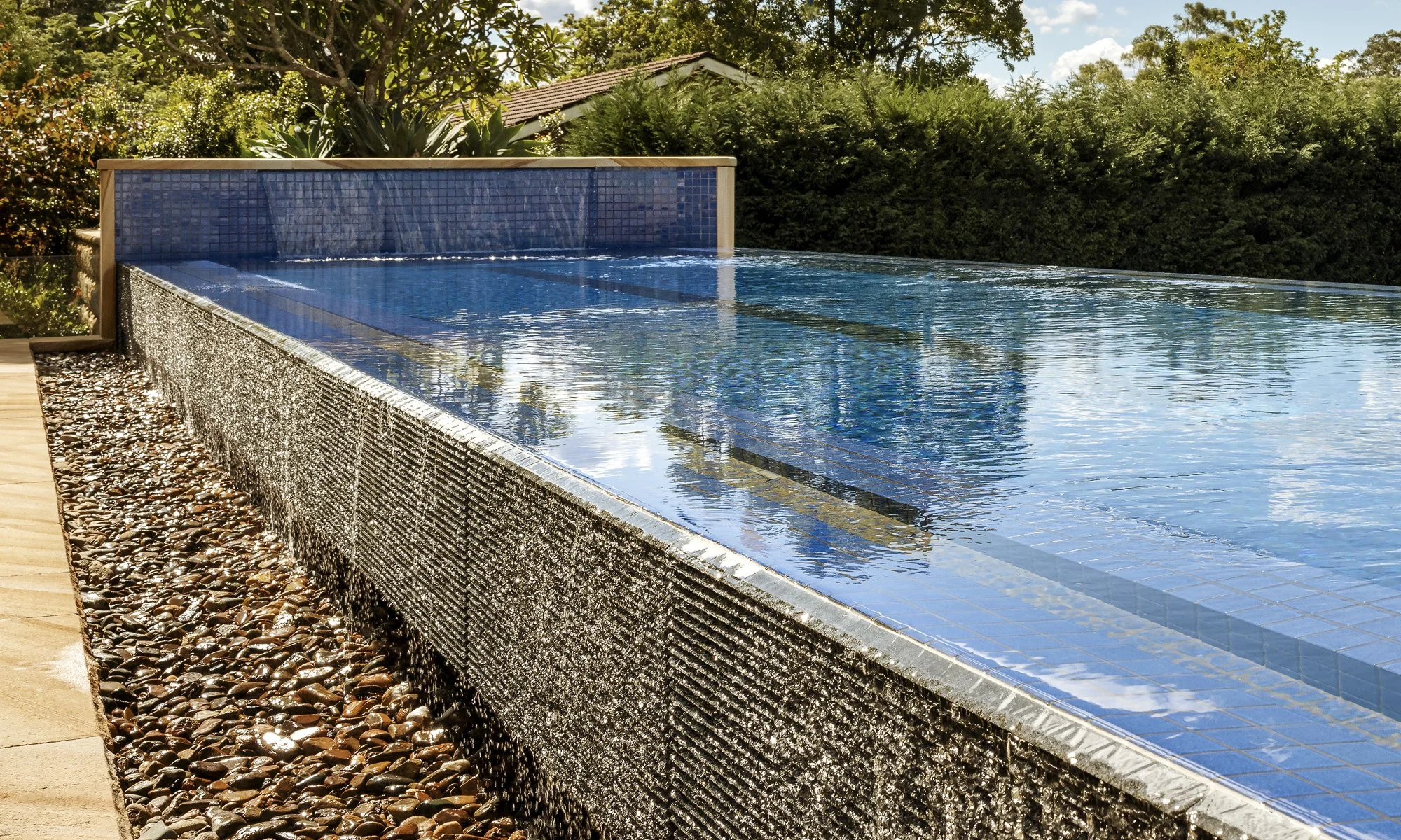 Outdoor infinity pool with water cascading over the edge, surrounded by lush greenery and trees.