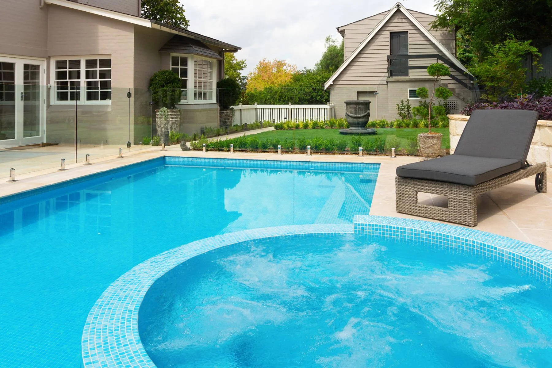 An outdoor pool area with a lounge chair on a stone patio, a small hot tub in the foreground, and a garden with a fountain and houses in the background.